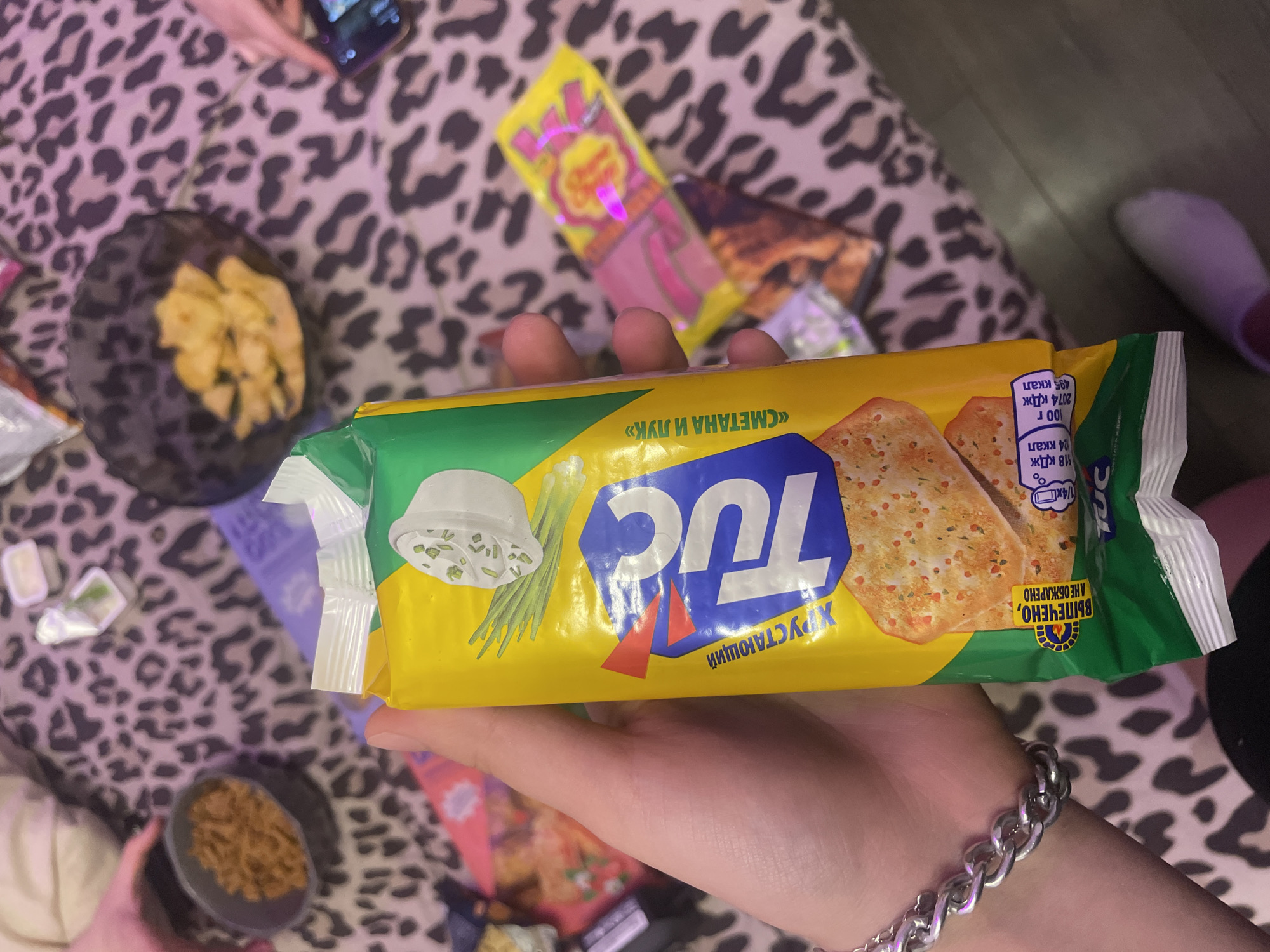 A hand, adorned with a chunky silver bracelet, holds a yellow and green package of TUC crackers. The crackers have a visible flavor of "Smetana and Onion," indicated by an image of sour cream and green onions on the packaging. The background is a table covered with a leopard-print tablecloth. Various items are scattered on the table, including bowls of snacks and a pink and yellow lollipop wrapper. A glimpse of a phone with a picture on the screen can be seen on the right. The scene appears to be a casual gathering, possibly a party or a get-together. The specific location of Zmeinyi, Russia is not discernable from the image.