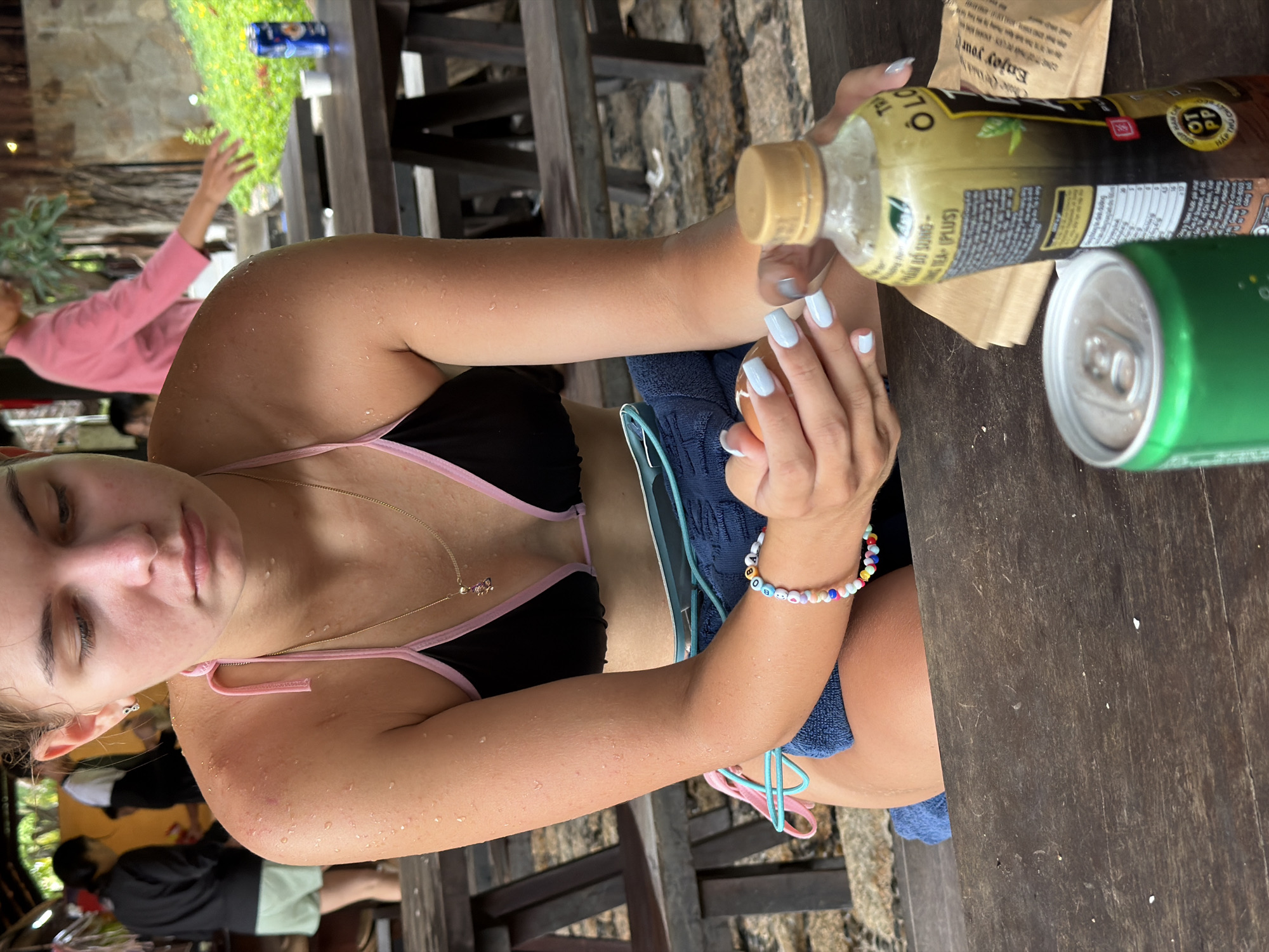 A young Caucasian woman is seated at a rustic wooden table outdoors, her face turned downwards and appearing contemplative. She is wearing a black and pink bikini top, and her skin is visibly wet with droplets, suggesting she has recently been swimming or exposed to water. A blue towel is draped over her lap. Her right hand is holding a piece of food, possibly an egg, and her nails are painted a light blue. She wears a colorful beaded bracelet on her left wrist that spells out "OBAMA".
In the foreground, a green soda can and a bottle of "Ô Long Tea Plus" are placed on the table. The bottle has Vietnamese text on its label. In the background, out of focus, other people are present, and lush green foliage and trees are visible, along with more wooden tables and benches. The scene appears to be in an open-air establishment, possibly a restaurant or a resort area in Nha Trang, Vietnam, given the presence of Vietnamese text on the beverage bottle and the tropical vegetation. The lighting suggests it is daytime.