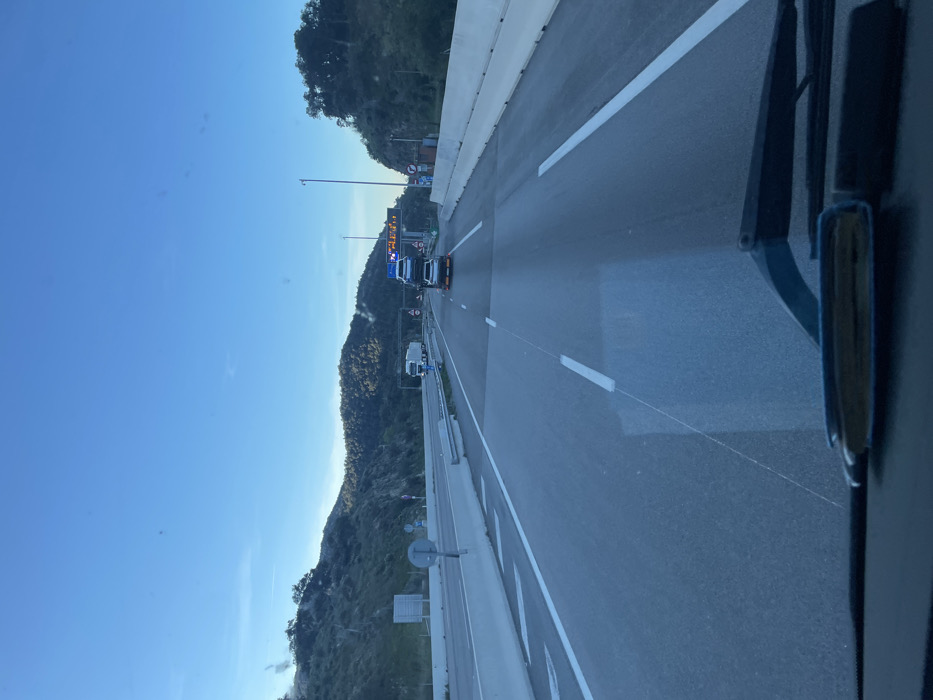 The image depicts a multi-lane highway environment near Le Perthus, France, viewed from within a vehicle. The sky is clear blue. Hilly terrain, densely covered with green vegetation and some rocky outcrops, flanks both sides of the highway.
In the immediate foreground, a vehicle's dashboard and two windshield wipers are visible. The asphalt road features white dashed and solid lane markings. White chevron markings are present on a merge lane to the left. Concrete barriers separate the main carriageway from the median and shoulders.
Vehicular traffic includes a white car carrier truck occupying the central lane, transporting four vehicles: two white cars are stacked on its upper deck, and two darker-colored vehicles are positioned on its lower deck. Further ahead in the left lane, a white semi-trailer truck is also in motion.
Overhead gantries with visible fixtures span the highway. On the right side, an electronic variable message sign displays illuminated yellow text, with the word "ALERT" clearly discernible. A small structure is situated roadside to the right. Distant mountainous terrain forms the horizon.