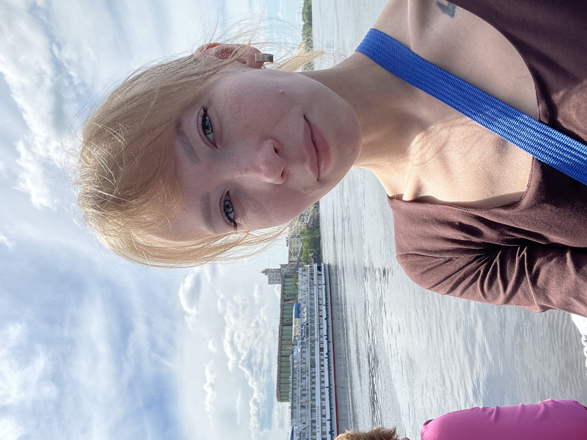 A young blonde woman is captured in a selfie taken outdoors on what appears to be a bright, partly cloudy day. She looks directly at the camera with a neutral to slightly smiling expression. Her blonde hair is parted and falls around her face, highlighting her light-colored eyes. She wears a brown top, and a bright blue strap runs diagonally across her chest, possibly from a bag. A small, dark tattoo is visible on her right shoulder, and a square-shaped earring is in her left ear.
The background reveals a wide body of water, likely a river, with gentle ripples reflecting the sky. Along the opposite bank, several buildings of a city are visible, suggesting an urban setting. Dominating the middle ground on the water is a large, white, multi-deck passenger ship. The name "Ф. И. ПАНФЕРОВ" (F. I. Panferov) is clearly visible in Cyrillic script on the side of the vessel. The sky above is bright with scattered white clouds. The image's tilted perspective indicates it is a selfie. In the bottom right corner, a portion of another person's head or shoulder, wearing a pink garment, is briefly seen. The scene depicts a river cruise or outing in Perm, Russia, given the location context and the name of the ship.