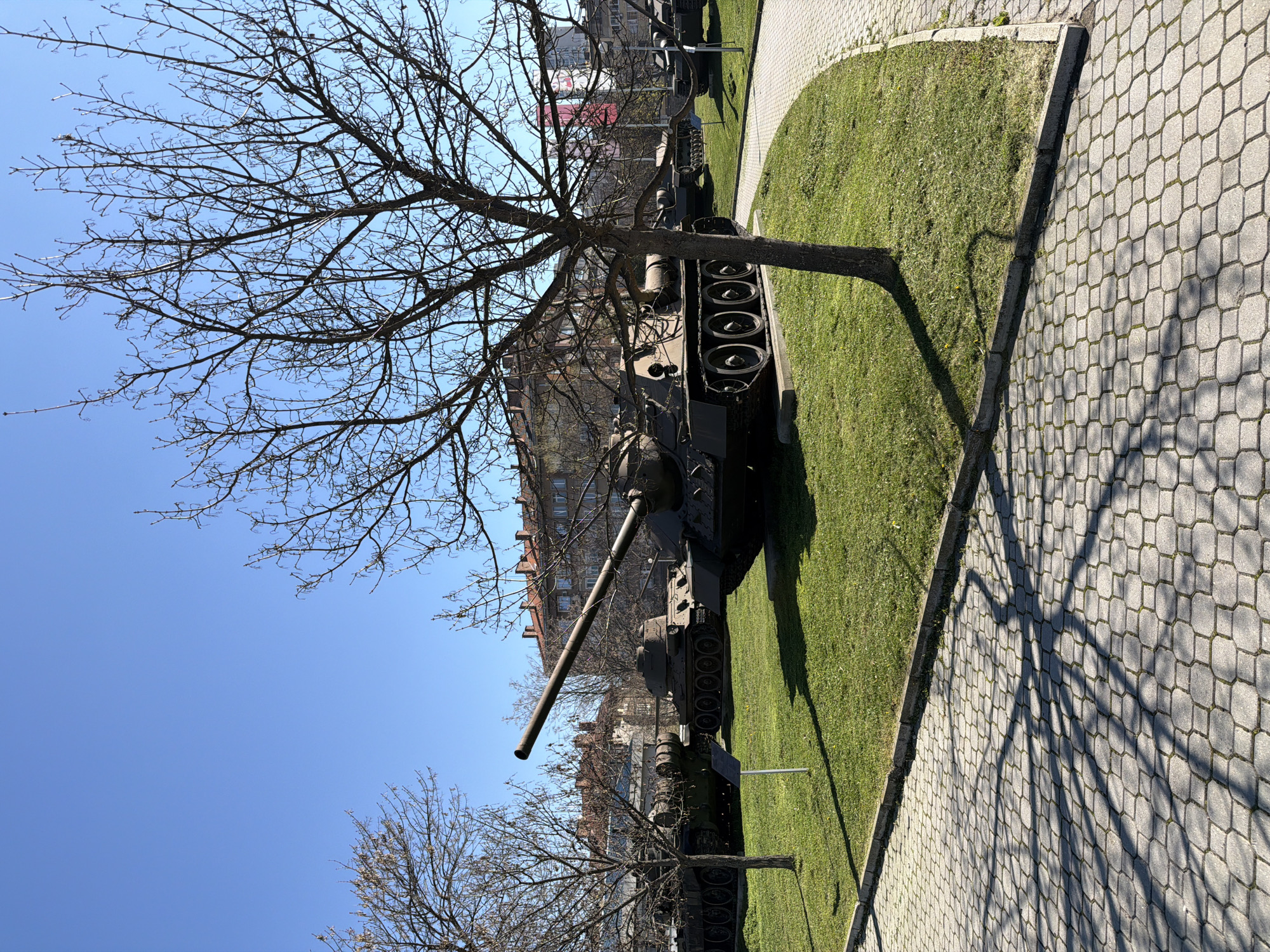 The image depicts a military tank on display in an urban setting. The tank is positioned on a patch of green grass next to a paved walkway. Bare tree branches frame the scene, with a clear blue sky in the background. Several buildings are visible behind the tank, suggesting a city location. The location is Unknown, Unknown.