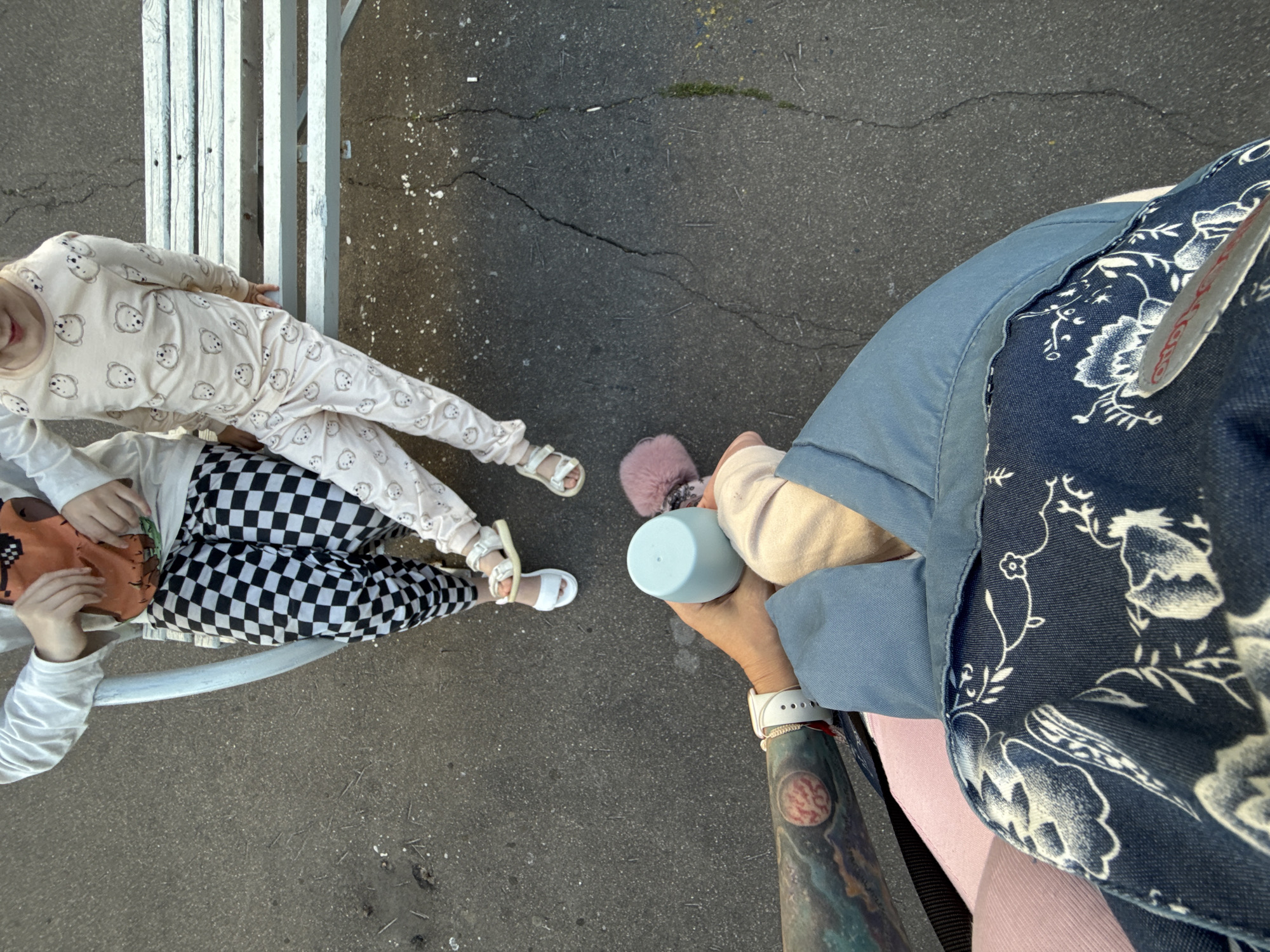 Two children are sitting on a white bench outdoors. The child on the left is wearing a white long-sleeved shirt with an orange graphic on the front and black and white checkered pants. They are wearing white sandals. The child on the right is wearing a light pink long-sleeved pajama set with a pattern of cartoon bears. They are also wearing white sandals. In the foreground, a person's arm with a tattoo is holding a light blue cup and a pink fluffy object. This person is wearing a blue garment with a floral pattern. The ground appears to be asphalt with some cracks. This scene is located in the city of Uzunovo, Russia.