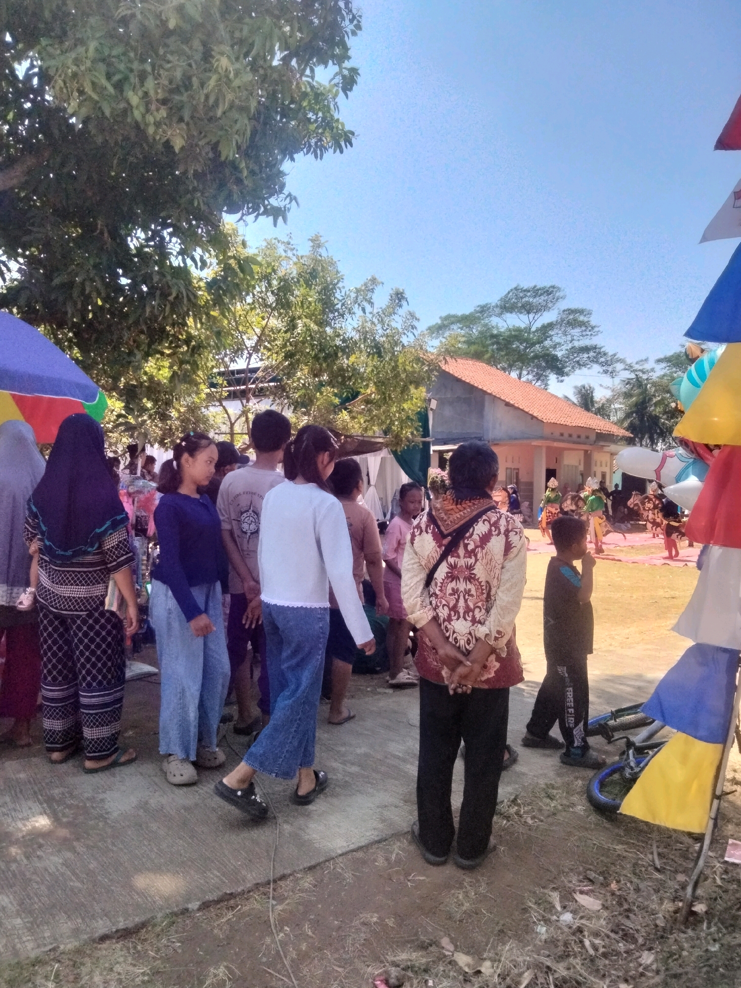 The image depicts an outdoor gathering, likely in the city of Cilacap, Indonesia. The scene is populated with several individuals of varying ages, engaged in casual observation and interaction. In the foreground, a group of women and children are present. One woman, facing away from the camera, wears a black and white patterned outfit and a dark blue hijab. Beside her, a young girl in a blue long-sleeved shirt and light blue jeans stands. To their right, another woman in a white long-sleeved shirt and blue jeans is also visible from behind. Further into the scene, a man in a light gray t-shirt and a boy in a dark shirt and pants are observed. The environment is characterized by a pathway or paved ground, with grassy areas and trees in the background. Behind the gathering, a building with a terracotta-tiled roof is visible, suggesting a residential or community setting. To the far right, colorful conical structures, possibly decorations or part of a stall, are present. A bicycle lies on the ground near these structures. In the mid-ground, a performance or event appears to be taking place on a raised area with a pink carpet, featuring individuals in traditional attire. The sky is clear and blue, indicating a sunny day. The overall impression is of a community event or a local festival.