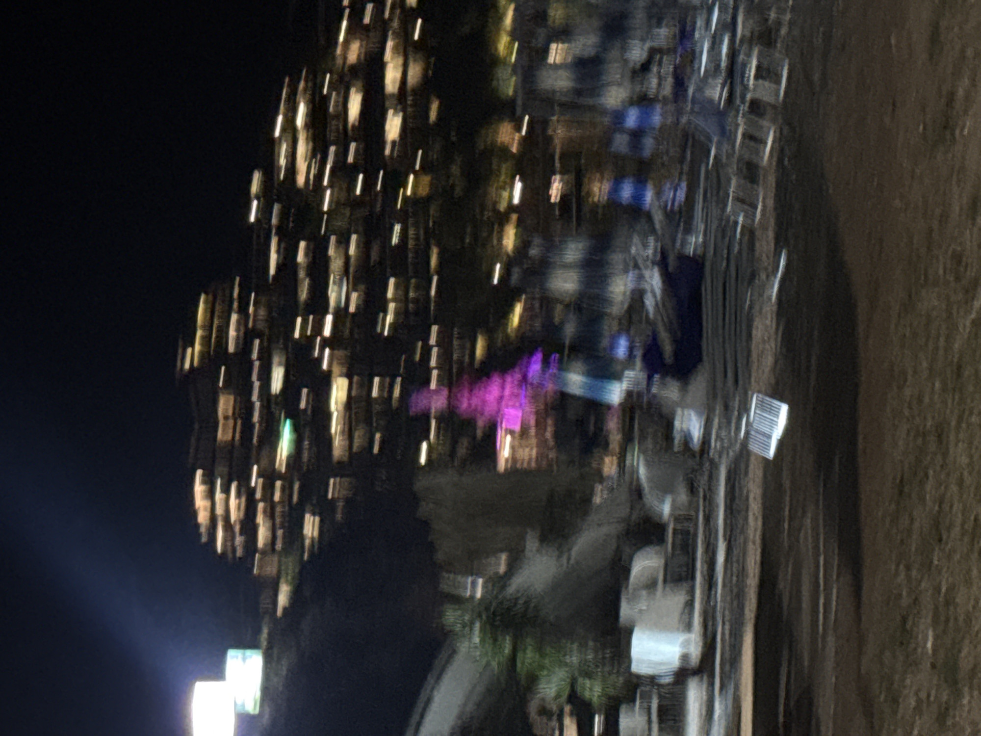 A blurry, nighttime photograph captures a hillside covered in illuminated buildings, likely hotels or apartments, in Budva, Montenegro. The lights create horizontal streaks across the dark sky. In the foreground, a beach or promenade area is visible with several lounge chairs and umbrellas, some appearing to be set up for the day. A bright pink, illuminated structure, possibly a decorative installation or a light display, stands out amidst the darker elements. A white rectangular object lies on the ground in the lower portion of the image. The overall impression is one of a lively, illuminated resort town at night.
