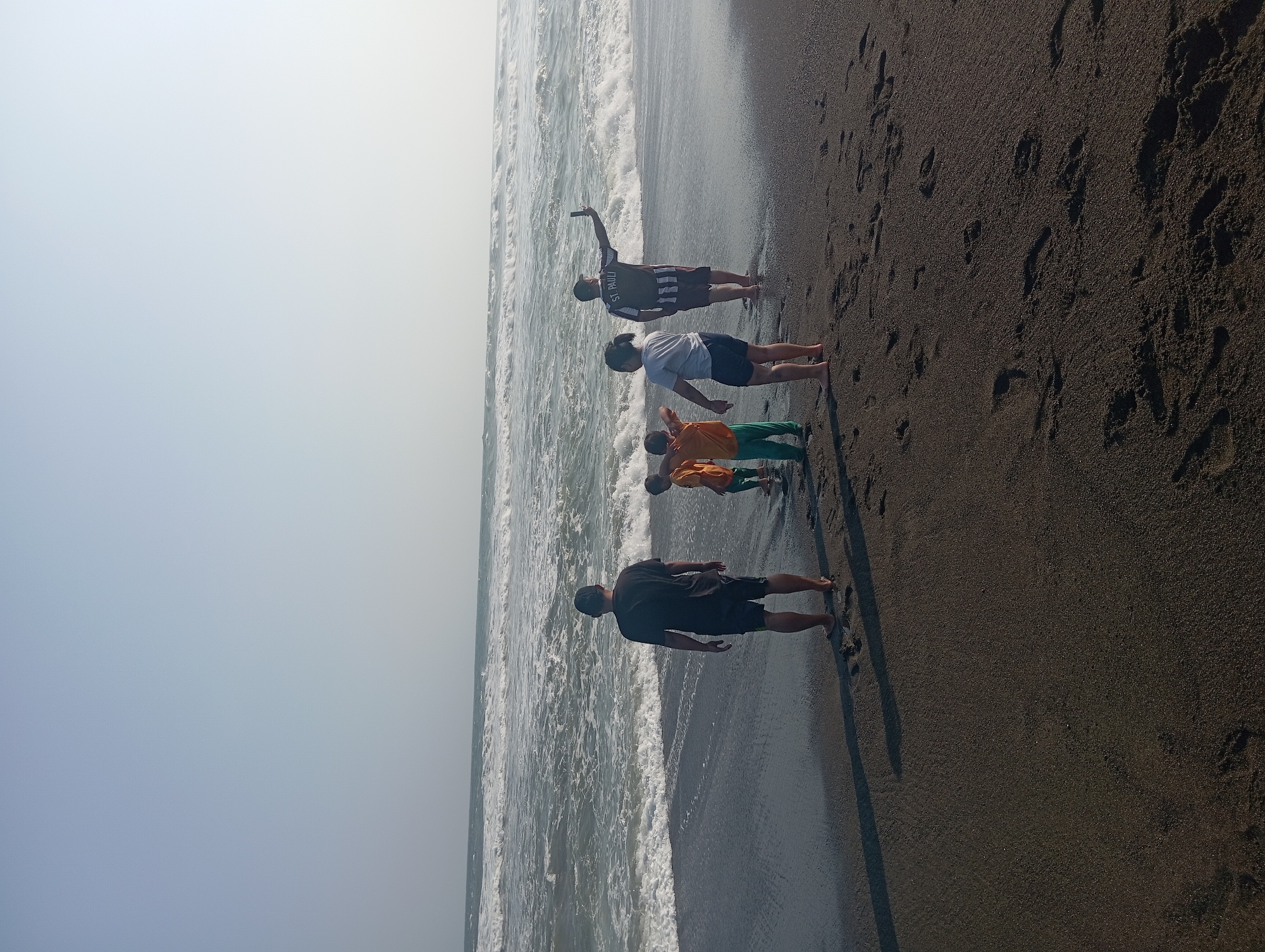This image depicts a family enjoying a day at Parangtritis Beach in Indonesia. The scene is set on the dark sandy shore with the ocean in the background. The water is rough with visible waves breaking onto the beach. In the foreground, a group of five individuals are present. A man in a dark t-shirt and shorts stands facing the ocean. To his right, three children are gathered. One child wears an orange shirt and green pants, another a yellow shirt, and a third a white t-shirt and dark shorts. Further to the right, a man in a striped shirt and shorts is taking a selfie with his phone. The sky is clear and bright, suggesting a sunny day. Footprints are visible in the sand, indicating recent activity on the beach. The overall atmosphere is one of leisure and recreation.