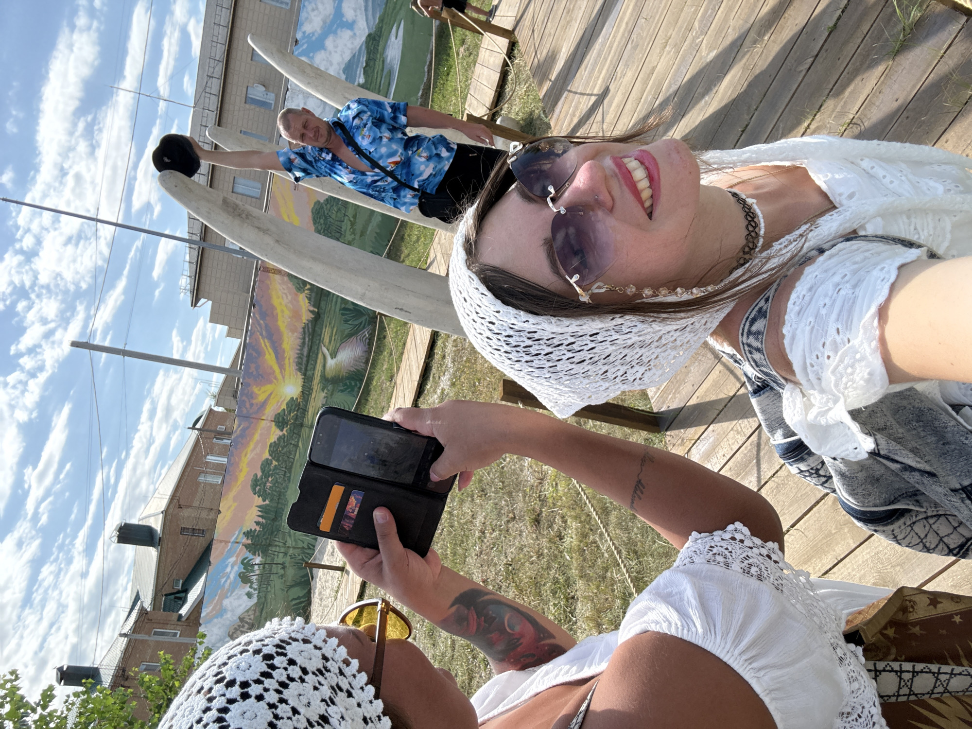 Two women are posing for a photo with a man in the background. One woman in the foreground, wearing a white headscarf, white top, and sunglasses, smiles at the camera. The other woman, also wearing a white headscarf and white top, is taking a photo of the group with her phone. The man in the background, wearing a blue floral shirt and black pants, holds a black hat above his head. They are on a wooden boardwalk, with a mural of a landscape visible behind them. The scene appears to be outdoors in Sol-Iletsk, Russia, with buildings and a cloudy sky in the distance.