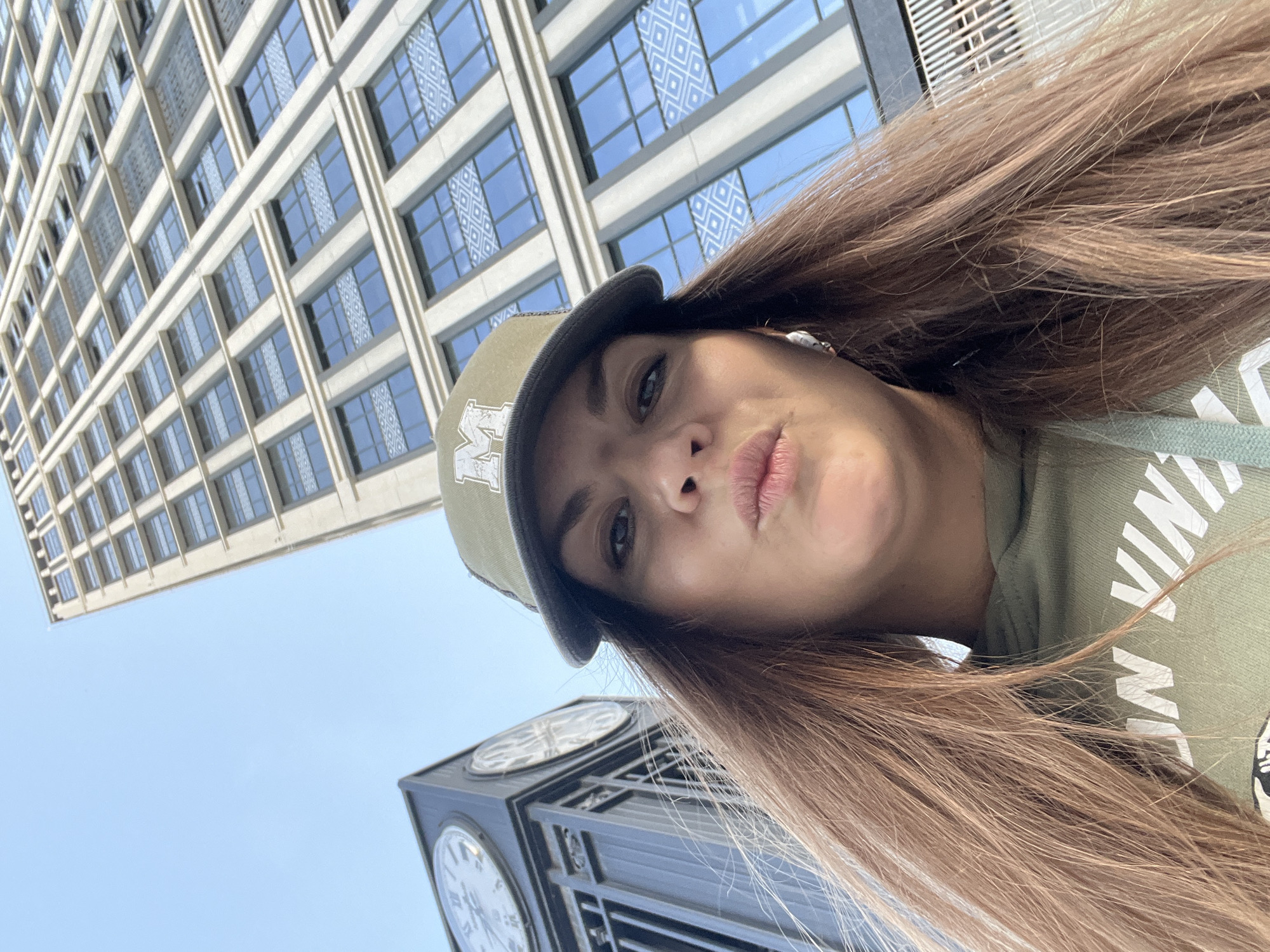 A selfie of a woman wearing a green cap and sweatshirt with long brown hair is presented against an urban backdrop. The image is taken from a low angle, capturing a tall building with numerous windows to the left of the subject and a clock tower partially visible at the bottom. The sky appears pale blue. There is insufficient information to place the location within Novosibirsk, Russia, but there are buildings and clock towers present, therefore the urban background matches Novosibirsk.