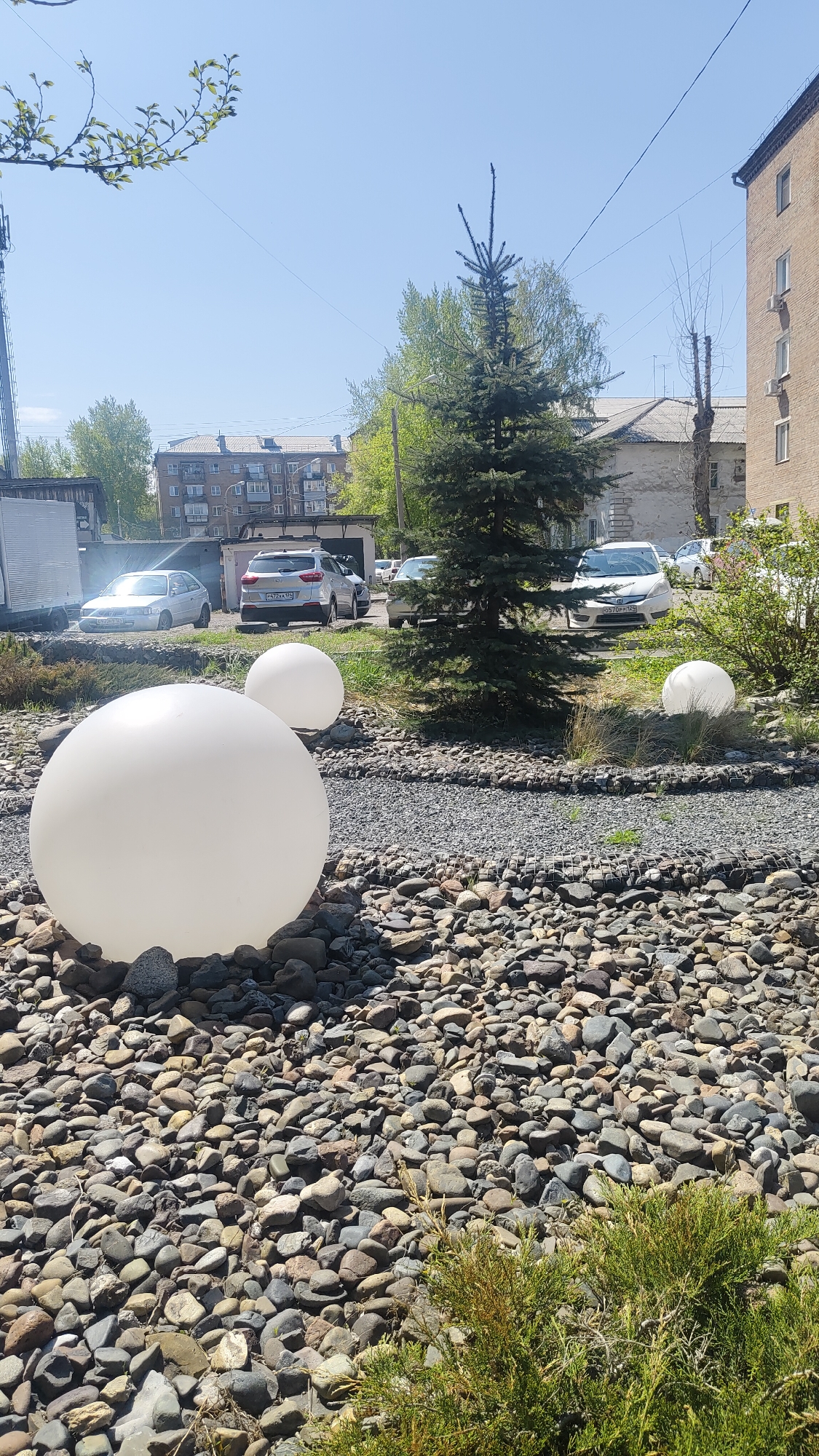 The image depicts an outdoor scene in what appears to be Krasnoyarsk, Russia, based on license plates. The foreground is dominated by a ground cover of varied-sized river rocks or pebbles, among which are three large, white spherical objects, possibly decorative lights. A low-lying evergreen shrub occupies the bottom right corner.
Behind the rocks, a small evergreen tree stands in a slightly elevated area filled with smaller, crushed rock material. Several vehicles, including a white SUV with license plate "P472XA 124" and another white car with license plate "O570PP 124", are parked in the background, partially obscured by the tree. A white van is visible on the left side of the frame.
Buildings typical of Russian urban architecture are seen in the background under a clear, blue sky. Overhead power lines stretch across the top of the frame. The image captures a mix of natural and urban elements within a public or semi-public space.