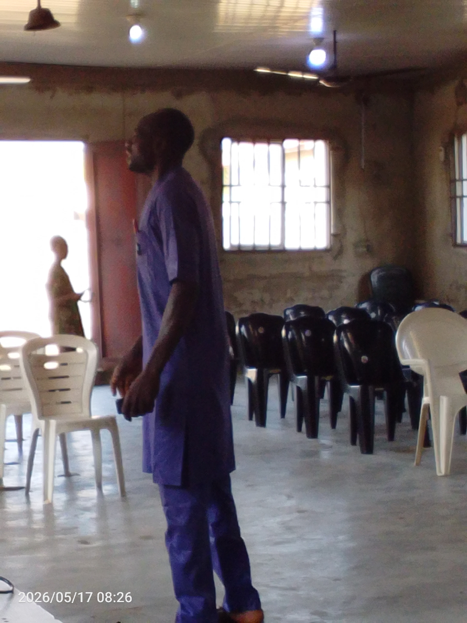 **Location:** Interior space, Lokoja, Nigeria.
**Scene:** A man dressed in a purple outfit is standing in a room with rows of chairs. There are two windows with metal bars behind him. Another person appears to be entering through a doorway in the background.
**Objects:** Chairs (both white and black), windows with metal bars, a door, and light fixtures on the ceiling.
**People:** Two individuals are visible in the frame. One is standing in the foreground, and the other is entering the doorway.
**Date/Time:** The timestamp at the bottom left corner suggests the image was taken on May 17, 2026, at 08:26.
