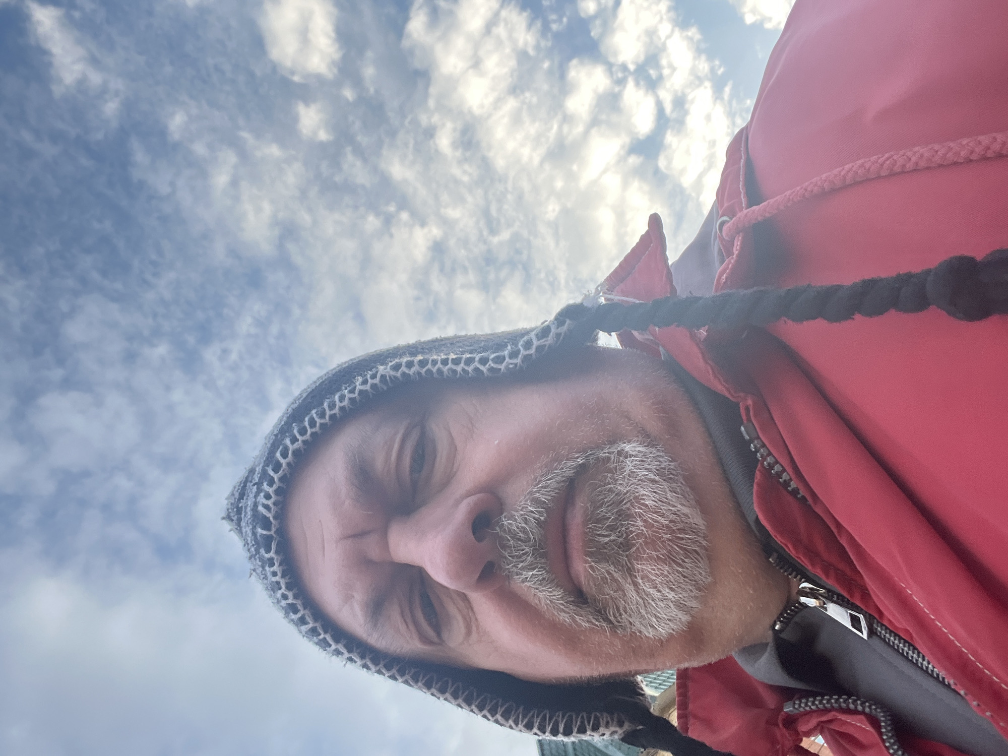 A low-angle, close-up self-portrait features a Caucasian male with a gray and white goatee, wearing a knit hat with a braided element and a red jacket. The man's face is the primary focus, and he appears to be looking downward towards the camera. The background shows a cloudy sky. There is no indication of Podolsk, Russia, or any other specific location. The image does not contain enough information to infer any actions or interactions beyond the act of taking a selfie.
