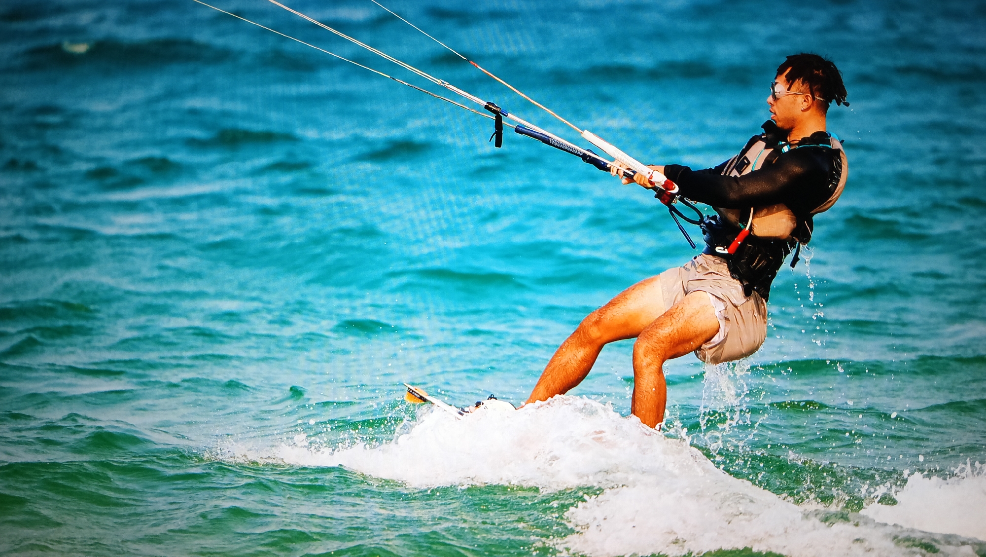 A person is kitesurfing on the ocean. The person is wearing a wetsuit jacket, life vest, sunglasses, and gray shorts. The kitesurfing board is yellow and white. The ocean is turquoise and green. The location is likely Nha Trang Ward, Vietnam, given the provided context.