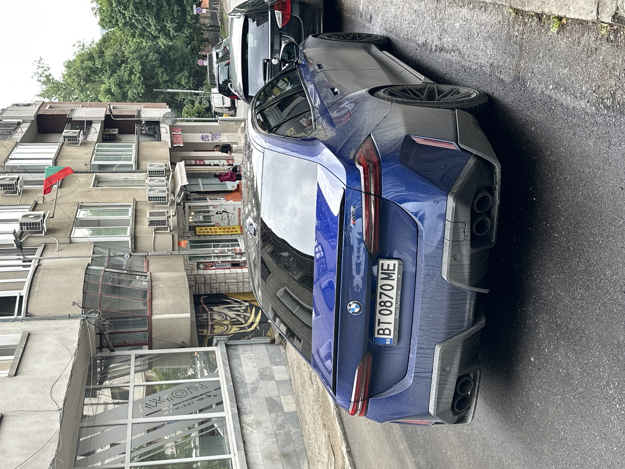 The image captures a street scene in Veliko Tarnovo, Bulgaria. A blue BMW with license plate "BT 0870 ME" is parked next to a dark colored SUV, partially obstructing the view. To the left, a multi-story building with numerous air conditioning units affixed to its facade is visible. A Bulgarian flag hangs from one of the windows. Below, a shop front with the sign "AXIOM" is seen. On the building, a mural with a black and yellow design is visible. People are present on the street and balconies, engaged in unclear activities. The road is wet, suggesting recent rainfall. The overall atmosphere is urban and lived-in.