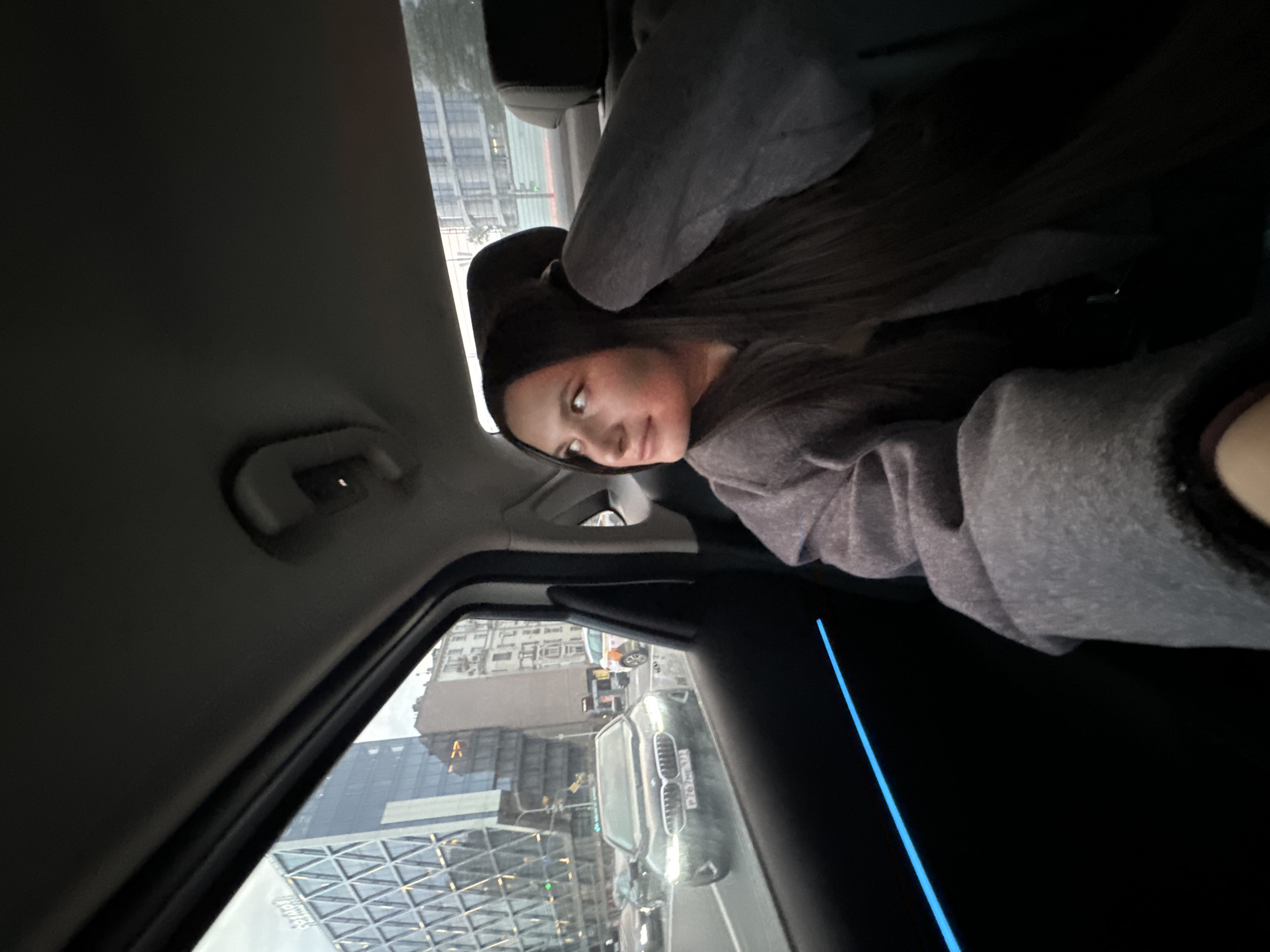 A woman with dark hair and wearing a gray hooded sweatshirt sits in the passenger seat of a car, looking towards the camera. The car's interior is visible, including the dashboard and a blue accent light. Through the car window, a city scene is visible, with buildings including the "Aquamarine III" business center on the left side. Other vehicles are present on the road. Based on the architecture and street layout, the location is likely in the city of Moscow, Russia.