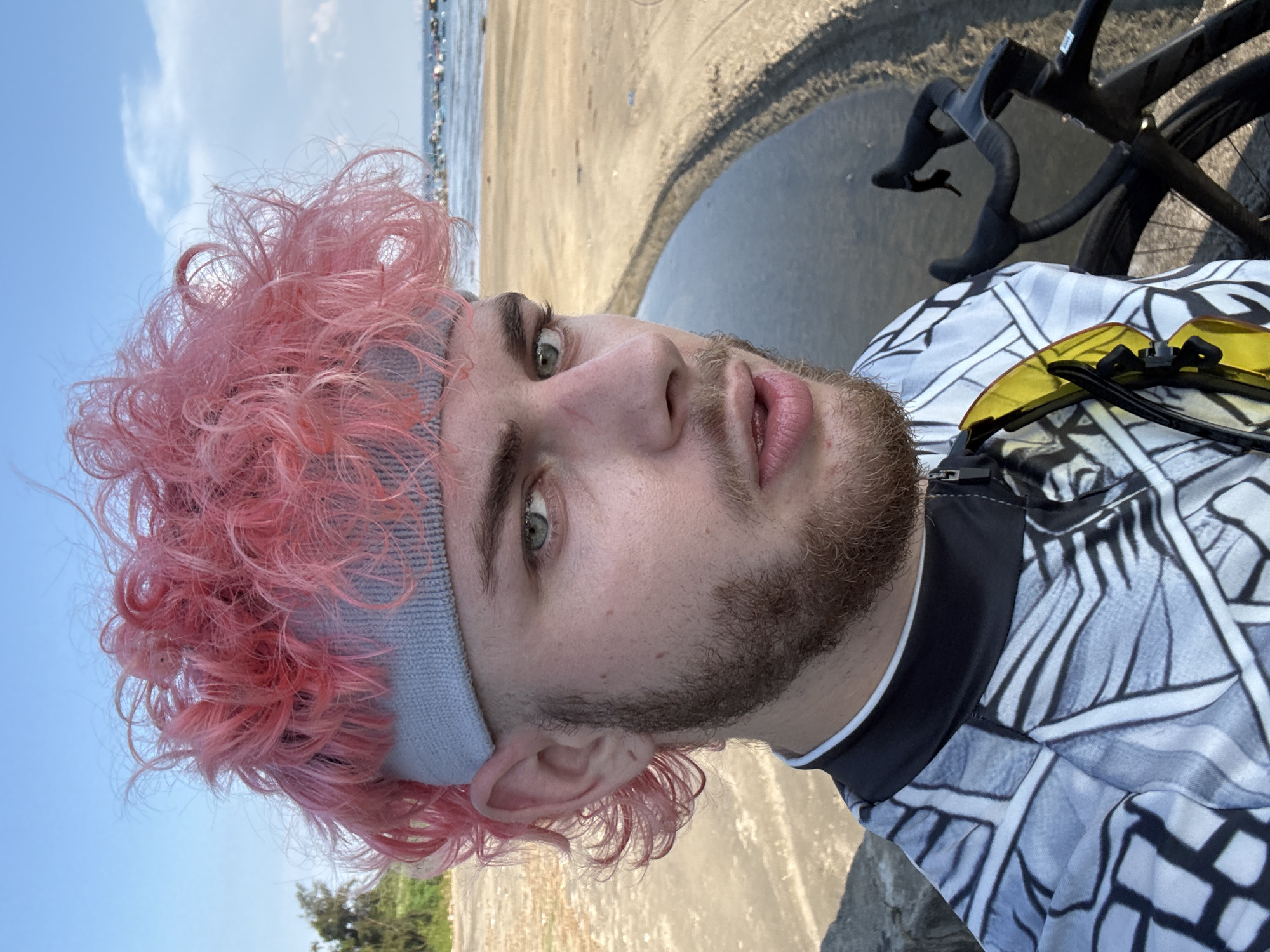 A selfie shot shows a light-skinned man with pink, curly hair and a grey headband. He has green eyes, a beard, and is wearing a patterned cycling jersey. Yellow sunglasses are attached to the jersey. In the background, a black bicycle is visible, partially cut off in the frame. The location appears to be a beach, with sand and water visible. Given that the location should be in Da Nang, Vietnam, the photo could potentially be taken along My Khe Beach.