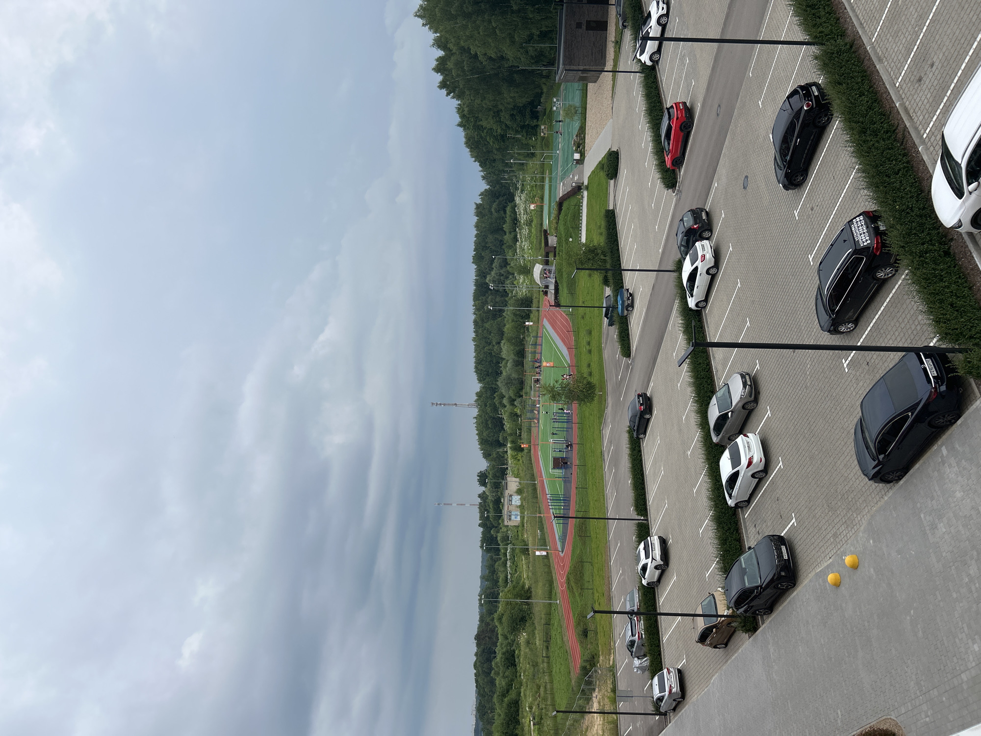 The image depicts an outdoor parking lot in the city of Kaluga, Russia, with a sports complex in the background. The sky is overcast and gray. The parking lot is paved with interlocking gray pavers and marked with white lines indicating parking spaces. There are numerous cars parked in the lot, including silver sedans, white SUVs, a red hatchback, black sedans, and a dark blue pickup truck. A few people are visible in the distance at the sports complex, which includes a running track, a soccer field, and basketball courts. Trees and greenery surround the sports complex and extend to the horizon. A low-rise building, possibly part of the sports facility, is visible on the right side of the image. The overall scene is that of a public recreational area on a cloudy day.
