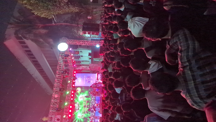 The image depicts an outdoor night concert or event in Dhaka, Bangladesh, with a large, dense crowd of individuals, predominantly young adults, facing a brightly lit stage. The audience, seen mostly from behind, is standing shoulder-to-shoulder under artificial stage lighting that casts red, green, and blue hues.
On the elevated stage, a musical band consisting of approximately five to seven members is actively performing with instruments such as guitars and possibly drums or keyboards. Two prominent LED screens form the stage backdrop, displaying a green logo with text on the left and a blue-toned graphic with text on the right. The stage structure comprises metal scaffolding, supporting numerous spotlights and wash lights that illuminate the performers and the immediate area. Large speaker stacks are visible on either side of the stage. A bright, spherical white light fixture is mounted on the scaffolding to the right of the stage.
In the background, behind and to the left of the stage, a multi-story building with a facade of rectangular windows is visible. The architecture suggests an institutional or public building. To the right of the stage and building, a tall palm tree and other dense green foliage are present. The sky above is dark, indicating nighttime, with some atmospheric particles or moisture illuminated by the intense stage lights.