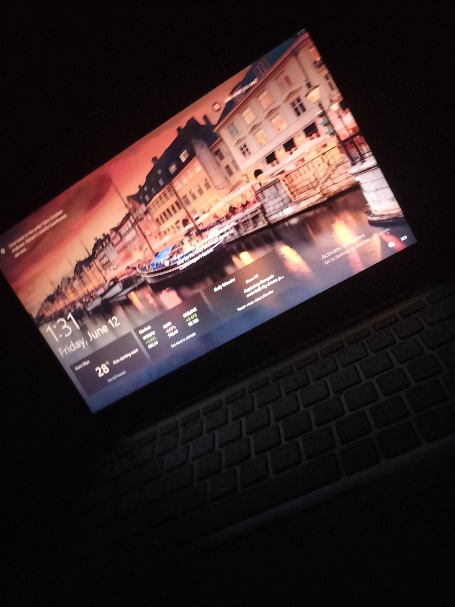 The image shows a laptop screen displaying a Windows lock screen. The background is a picturesque scene of a canal lined with buildings, likely in Europe. The time is 1:31 on Friday, June 12. There is weather information showing a temperature of 28 degrees Celsius and a forecast of rain starting soon. Stock market data is also visible, with USD INR and USD MYR listed. In the lower right corner, there is a prompt to "Activate Windows." The laptop is placed on a keyboard. The scene is dimly lit, suggesting it's nighttime or a dark room. There are no people or animals visible in the image. There is no indication of the image being related to Jalingo, Nigeria; all visible information points to a Western setting.
