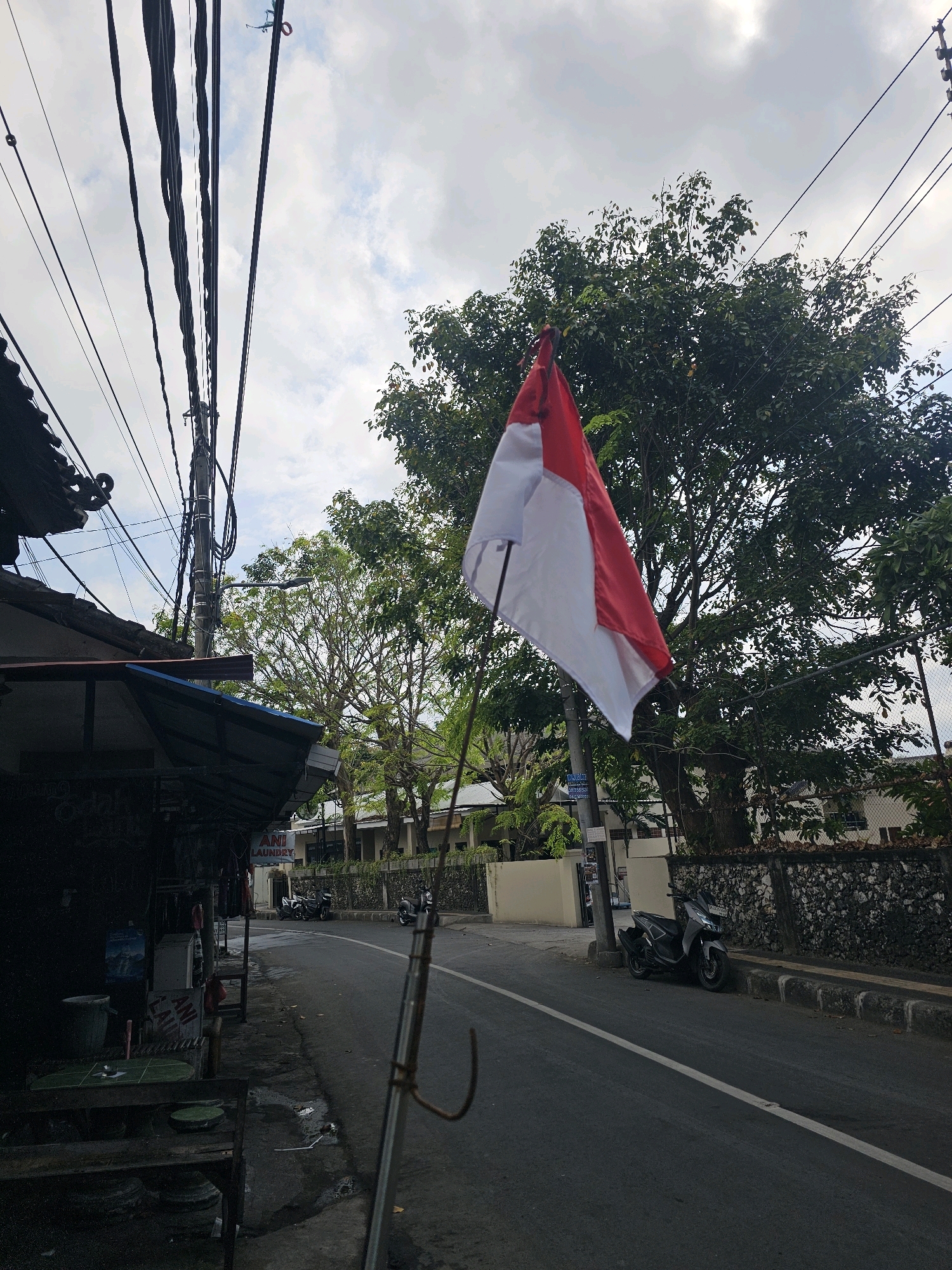 The image captures a street scene in Denpasar, Indonesia, dominated by an Indonesian flag waving in the wind. The flag, with its red and white colors, is prominently displayed in the foreground, slightly off-center to the right. Behind the flag, a large, leafy green tree provides a natural backdrop. The street itself is paved and appears to be a two-lane road.
On the left side of the street, there is a row of buildings with awnings, suggesting commercial establishments. One of these buildings has a sign that reads "ANI LAUNDRY". A few motorbikes are parked along the side of the road. The sky above is overcast with some patches of blue peeking through, indicating a cloudy day. Electrical wires are visible crisscrossing the sky, supported by utility poles. The overall impression is of a typical urban street in a tropical climate. The foreground also shows a metal pole with a hook attached, possibly part of a temporary flag stand or a utility fixture. The scene is devoid of any visible people.