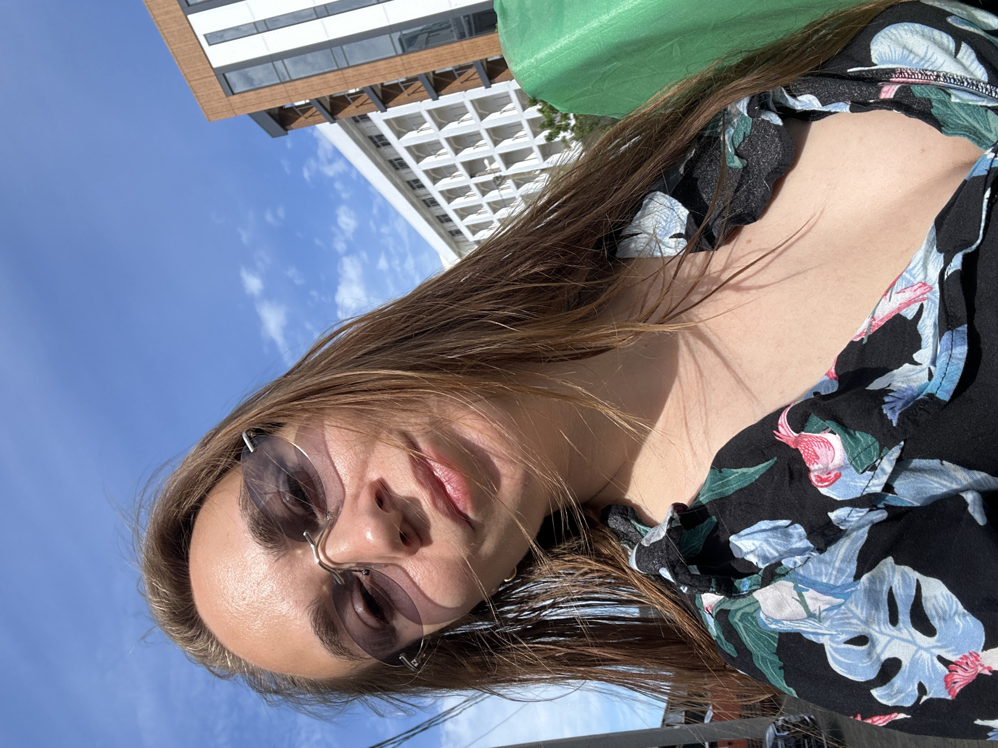 A woman with shoulder-length brown hair, wearing rimless sunglasses and a black floral-print blouse, stands outdoors on a sunny day. Behind her, a modern building with white balconies and a building with a brown facade are visible against a blue sky with scattered clouds. A green object, possibly a tarp, is to the right of the woman. The scene appears to be located in Vityazevo, Russia, a resort town on the Black Sea.