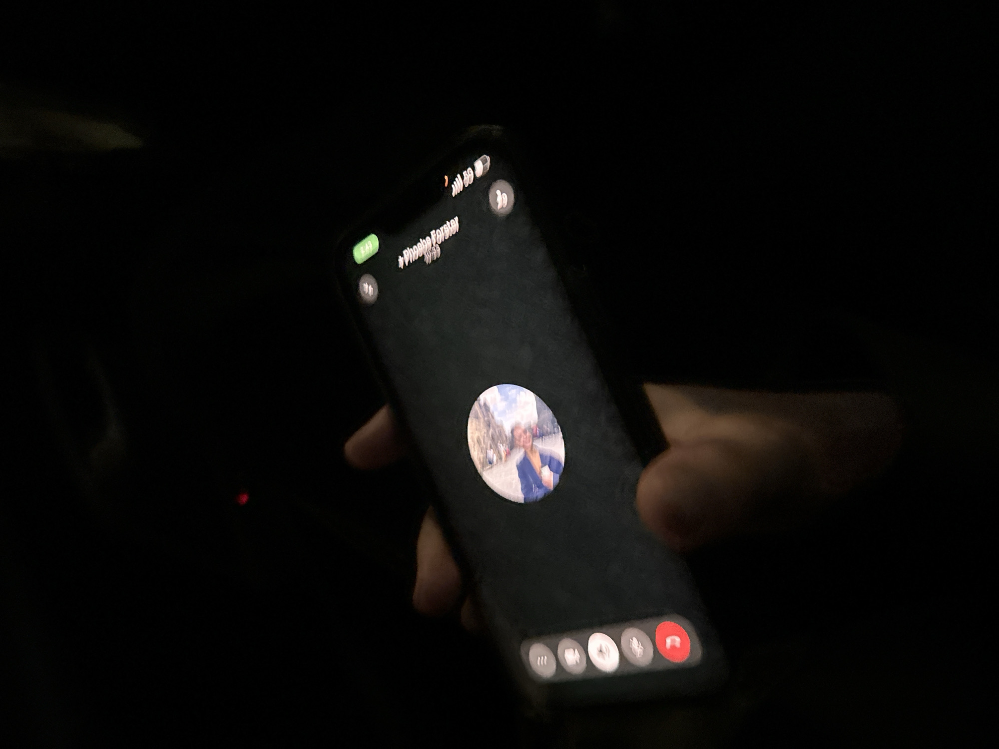 A person is holding a smartphone in their hand. The phone screen shows an active video call with a contact named "*Phoebe Forster*". The profile picture displayed on the call shows a person standing outdoors in a city with buildings and cloudy skies in the background. While the exact location is unclear, the architectural style suggests it could be a European city. However, based on the hint, I am going to say it could be a location in London, UK, but I can't confirm this without additional information. The phone is tilted at an angle, and the surrounding environment is dark, with only the light from the phone screen illuminating the hand and the device.