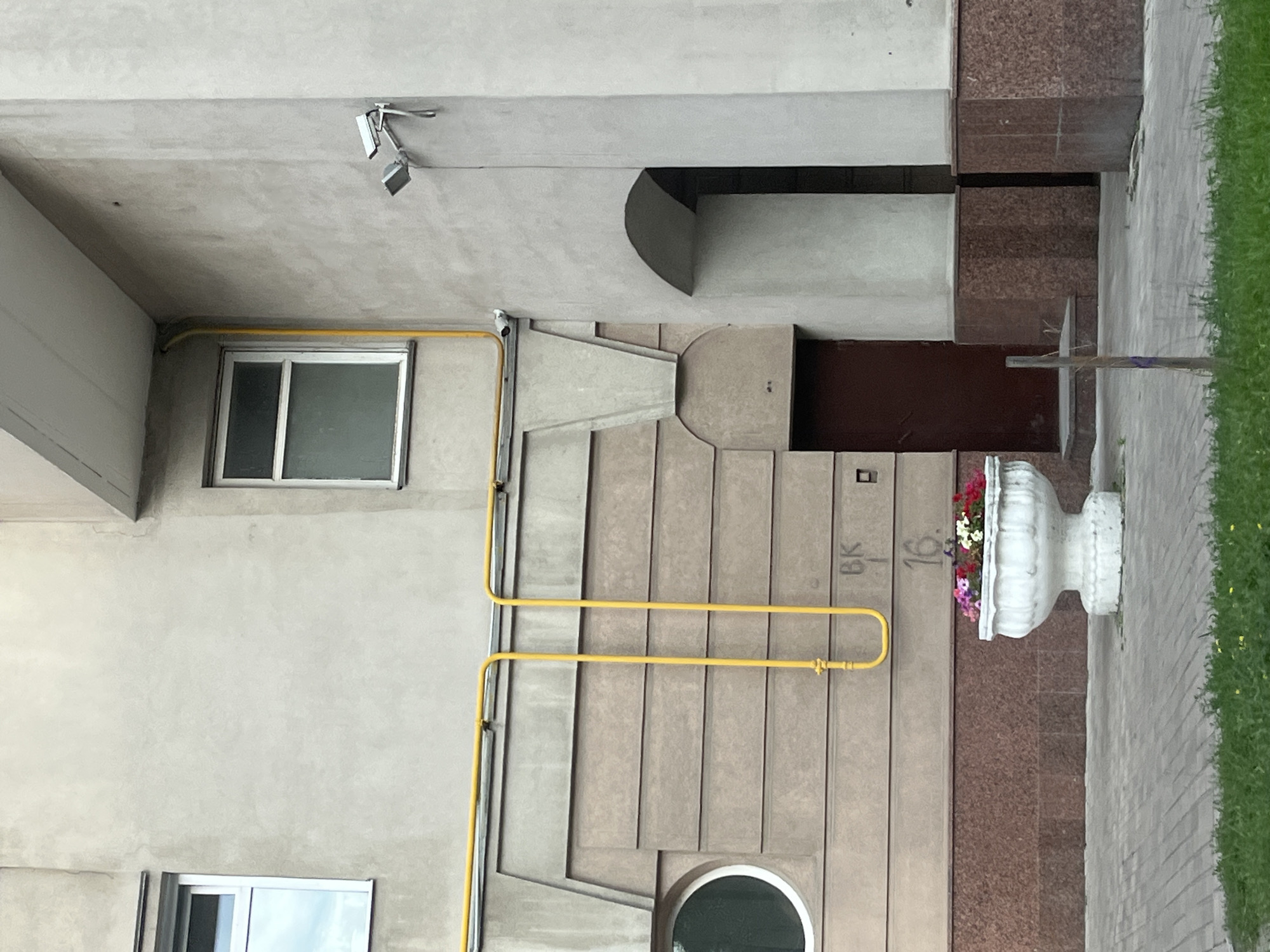 A medium shot captures the exterior of a light-colored apartment building in Novokuybyshevsk, Russia. The building features beige horizontal siding and a reddish-brown granite base. A bright yellow gas pipe runs horizontally across the facade and then vertically downwards. To the left, a circular window is visible. A large, white, ornate planter filled with colorful flowers sits on the paved walkway in front of the building. Above and to the right of the planter, scratched onto the siding, are the letters "BK" and the number "16". An arched doorway with a dark red door is situated to the right of the planter. Above the doorway, a security camera is mounted on the wall. The walkway is made of grey interlocking pavers, and a strip of green grass is visible in the foreground. A thin, wooden pole with a purple marker stands on the walkway.