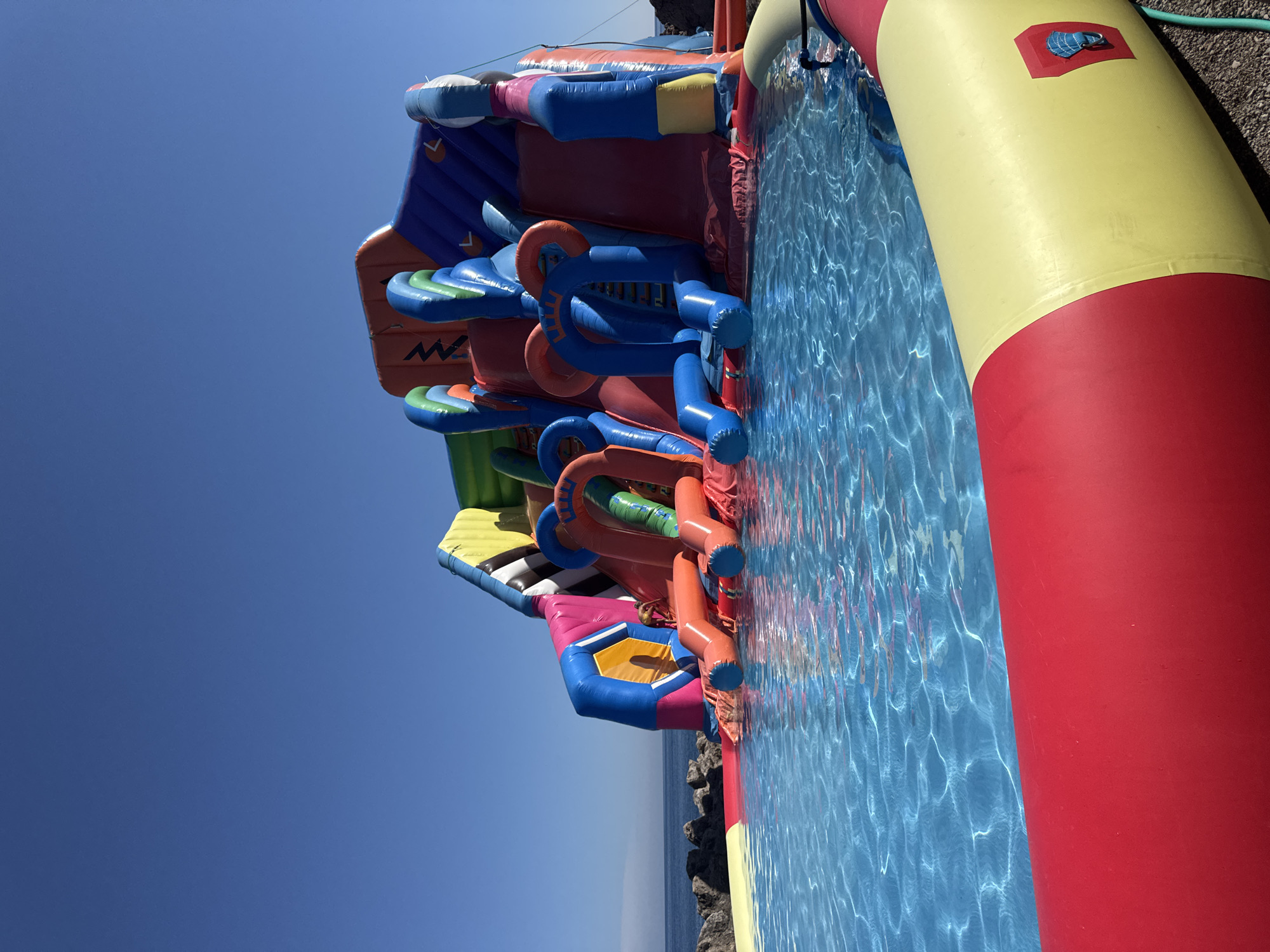 In Prijevor, Montenegro, on a clear, sunny day, an inflatable water park is set up near the sea. The park features a large, multi-colored structure with slides and climbing sections, appearing to be made of durable vinyl material. It is partially submerged in a bright blue, shallow pool of water, the surface of which is rippled and reflecting sunlight. The edge of the pool is defined by a thick, inflatable barrier with red and yellow sections. In the background, beyond the water park, rocky shoreline and the calm, blue sea are visible under a clear blue sky. No people are clearly depicted in the scene.