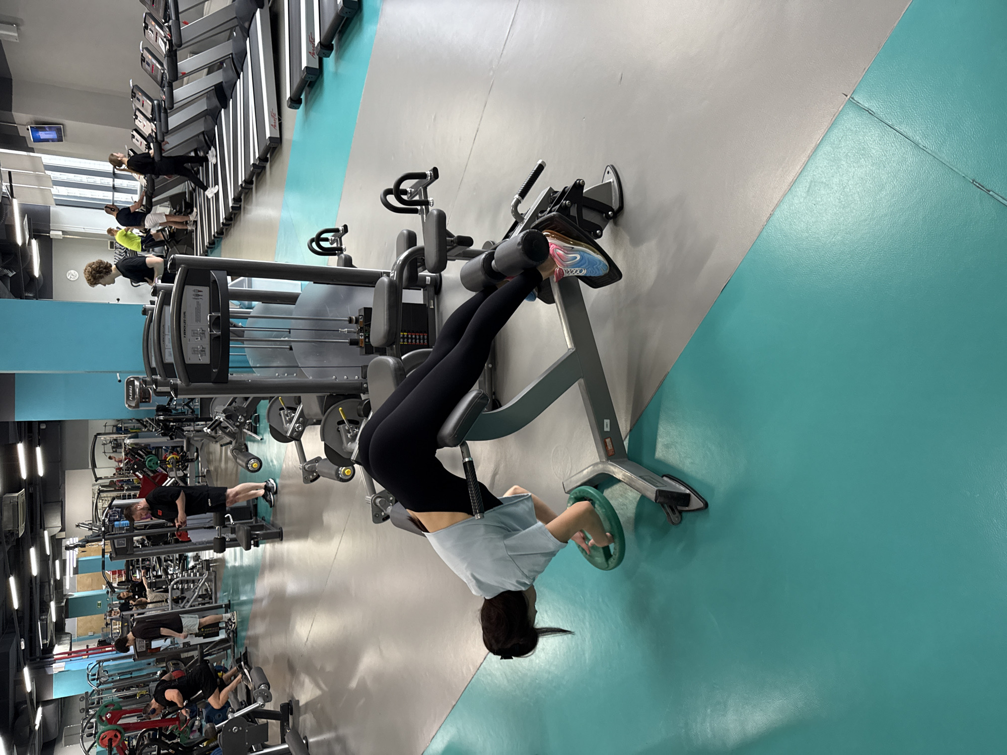 A woman in black leggings and a light blue t-shirt is using a leg press machine in a gym. The gym is modern with a two-tone gray and turquoise floor. Other gym equipment, including treadmills and weight machines, are visible in the background, with several people exercising on them. The location is a gym, which, from contextual knowledge, could be in Yekaterinburg, Russia.
