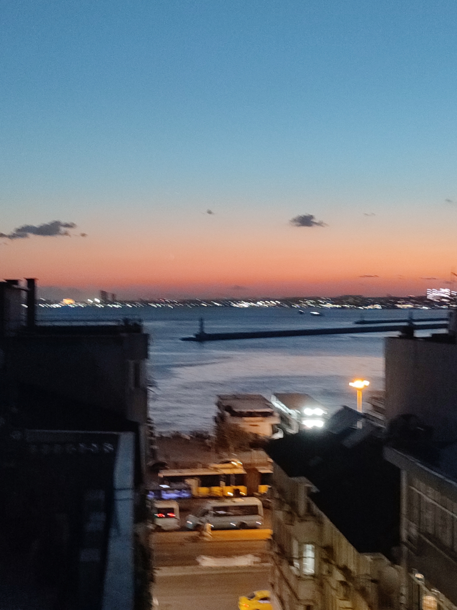 The image depicts a twilight scene overlooking the Bosphorus strait in Kadıköy, Turkey. The sky transitions from a deep blue at the top to hues of orange and pink near the horizon, with a few scattered clouds. Across the water, the lights of the cityscape are visible, including buildings and a lighthouse. In the foreground, the silhouettes of buildings dominate the left and right sides of the frame. The lower portion of the image shows a street scene with several vehicles, including buses and a yellow car. The overall impression is one of an urban environment at dusk, with a body of water separating the foreground from the distant city.