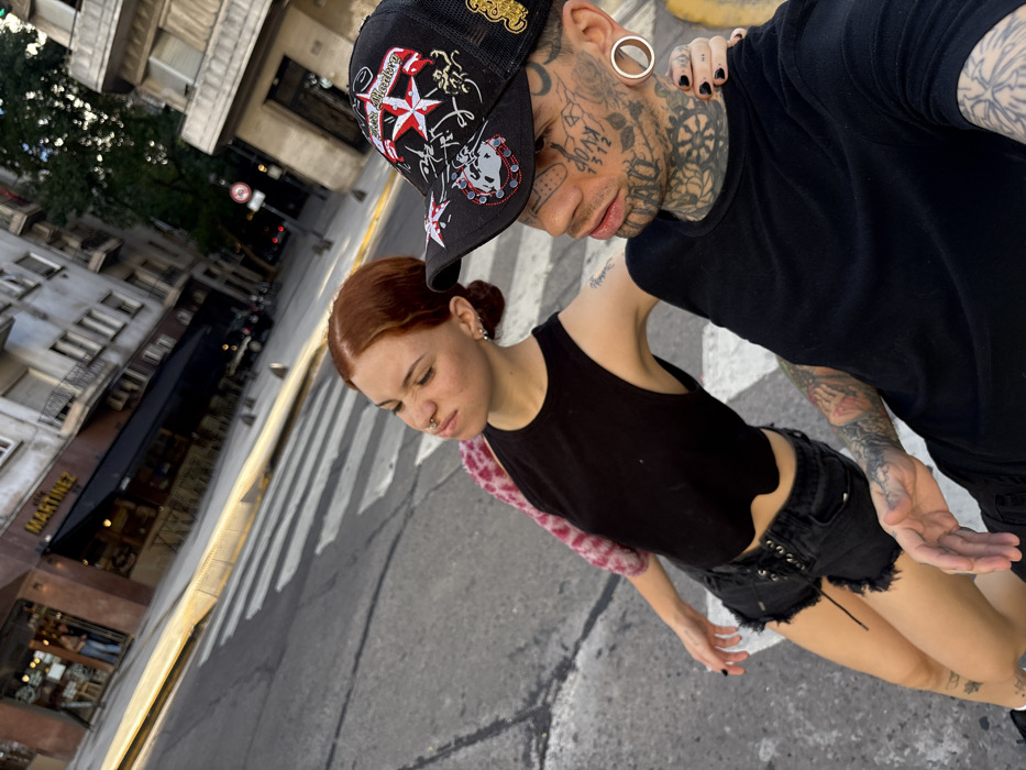 An urban street scene in Buenos Aires, Argentina, depicting two individuals interacting in the foreground. The female subject on the left has reddish-brown hair tied back, fair skin with light freckles, a septum piercing, and small hoop earrings. She wears a black sleeveless top with an irregular hem, black ripped denim shorts with front lacing, and a pink/red patterned garment visible over her left shoulder. An arm tattoo is present on her left bicep. Her left arm rests on the right shoulder of the male subject. The male subject on the right is heavily tattooed on his face, neck, and arms, and wears large, circular ear gauges. He wears a black mesh-back baseball cap adorned with multiple patches, including stars, text ("DIABLO" or similar), and other symbols in red, white, and gold. He wears a plain black t-shirt. His left arm is around the female subject's waist. Both individuals have dark nail polish on their visible hands. They appear to be posing for a close-up photo. The background features a multi-story urban building with an older architectural style, including a commercial establishment on the ground floor with a prominent gold "MARTINEZ" sign and a black awning. A street with white pedestrian crosswalk markings and yellow curbstones is visible, along with distant trees and other urban structures. The lighting suggests daytime, possibly late afternoon.