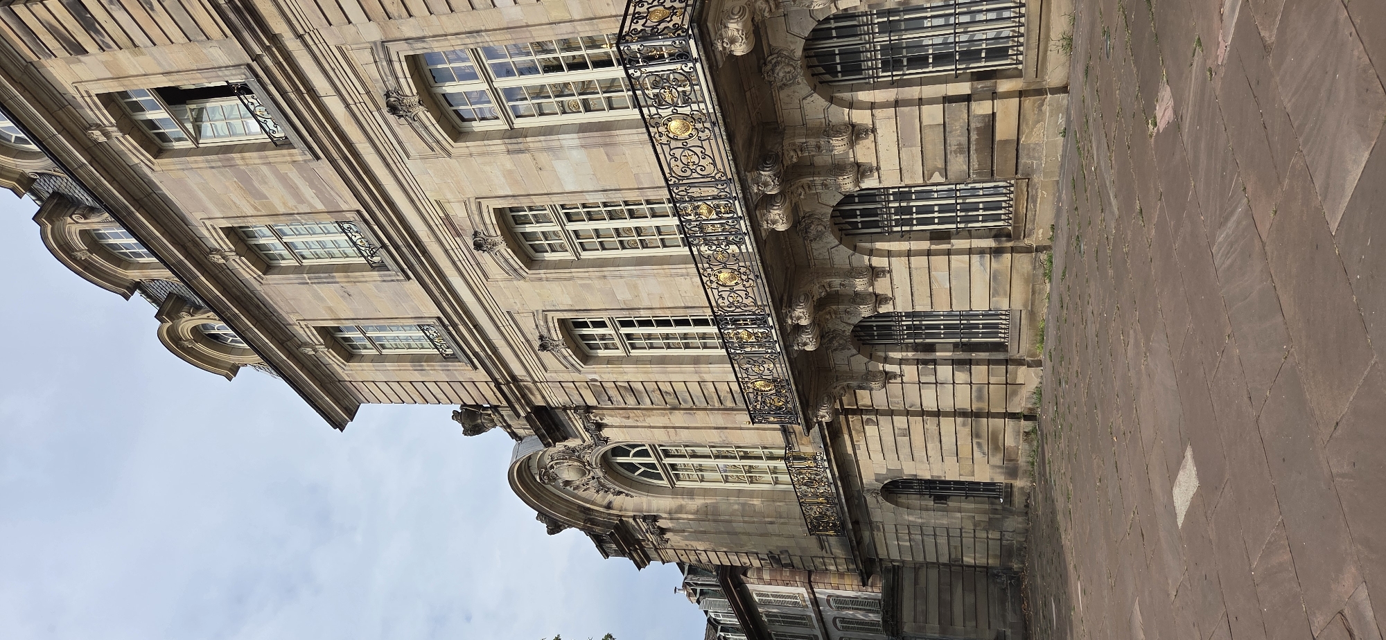 This is a full-shot, low-angle exterior photograph of a historic, ornate building made of light-colored sandstone, likely located in a European city. The building features a mansard roof with arched dormer windows. The facade is characterized by multiple stories with tall, rectangular windows, many with black iron window grates and some with open shutters. A prominent balcony with intricate black and gold metalwork runs across the upper floor, supported by decorative stone corbels. The ground level has arched windows with similar black grates. The paving stones in front of the building are large and flat, laid in a somewhat irregular pattern, with some grass growing in the cracks. The sky above is overcast with scattered clouds. The architectural style suggests a building from the Baroque or Neoclassical period.