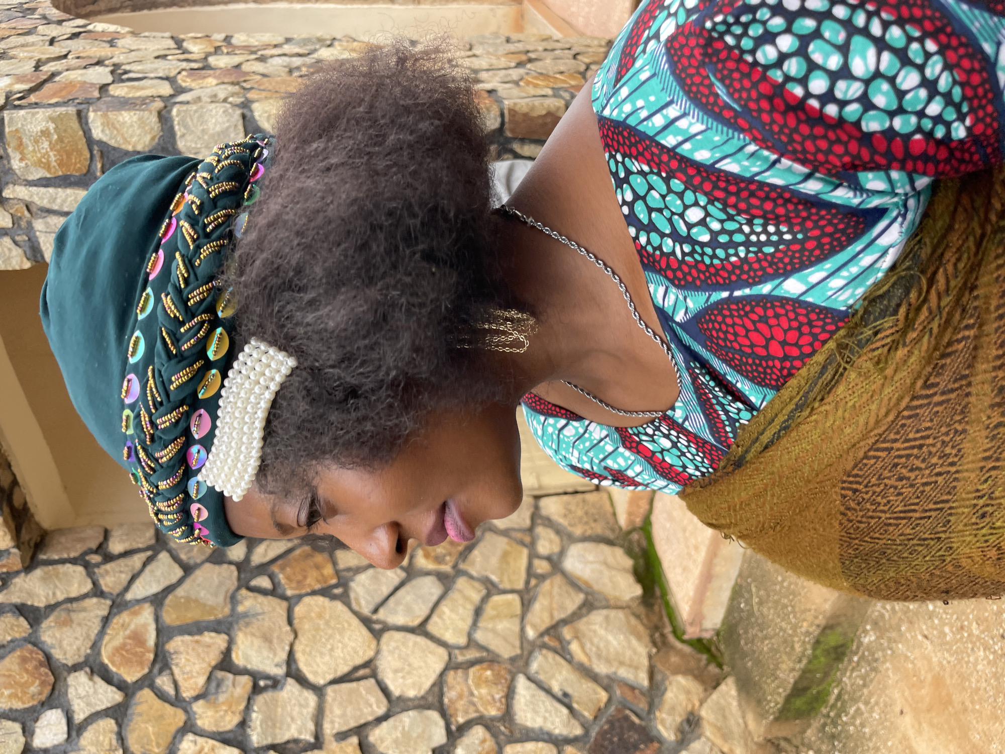 A woman is photographed from the side in an outdoor setting. She is wearing a green headscarf with colorful beaded embellishments, a pearl barrette, a patterned dress with red, teal, and white designs, a gold scarf draped over her shoulders, and a silver chain necklace. She also has gold chain earrings. The background consists of a textured stone wall and some steps. The lighting is natural and appears to be daylight. The scene suggests a cultural or artistic context. The specific location in Jos, Nigeria, is not discernible from the visual elements alone.