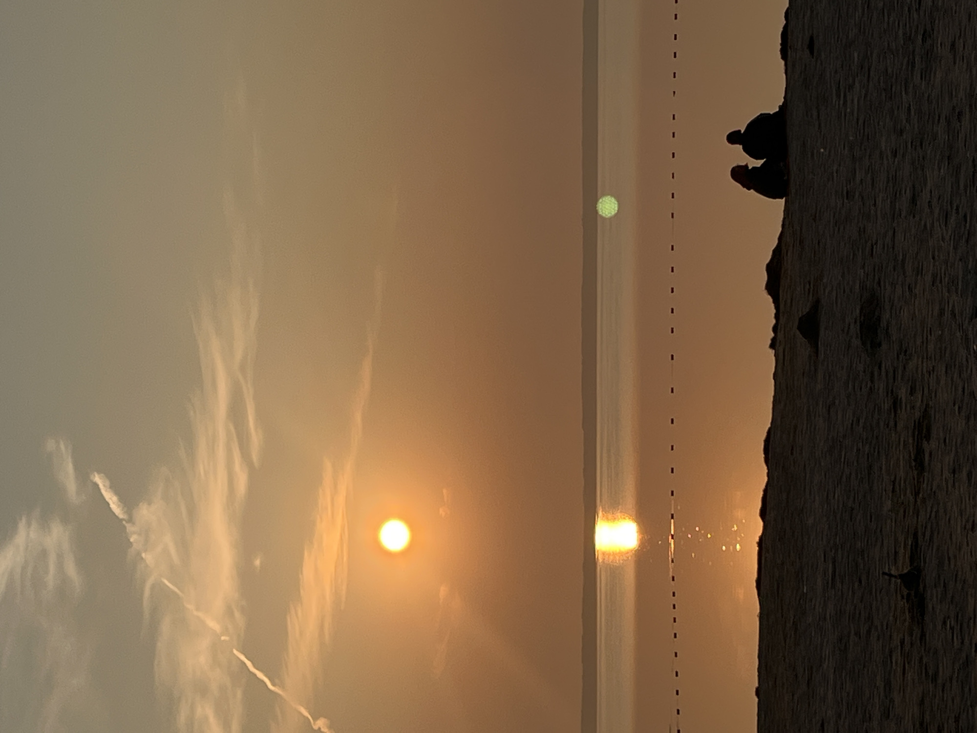 This image depicts a tranquil sunset scene on a beach, likely in Surakovo, Russia. The sun, a bright orange orb, is low in the sky, casting a warm glow across the horizon. Its reflection shimmers on the calm water of a large body of water, possibly a lake or the sea, which stretches to the horizon. A thin line of buoys or markers is visible on the water's surface, delineating a swimming area.
In the foreground, the sandy beach is dark, with the silhouettes of two individuals sitting together, facing the water and the sunset. They are dressed in casual clothing and appear to be enjoying the view in quiet companionship. A small, dark object, possibly a piece of driftwood or a toy, lies in the sand near them. The sky is a soft orange and yellow gradient, with wispy clouds adding texture. A faint contrail from an aircraft cuts across the upper left portion of the sky, a subtle reminder of the world beyond this serene moment. The overall atmosphere is one of peace and natural beauty.