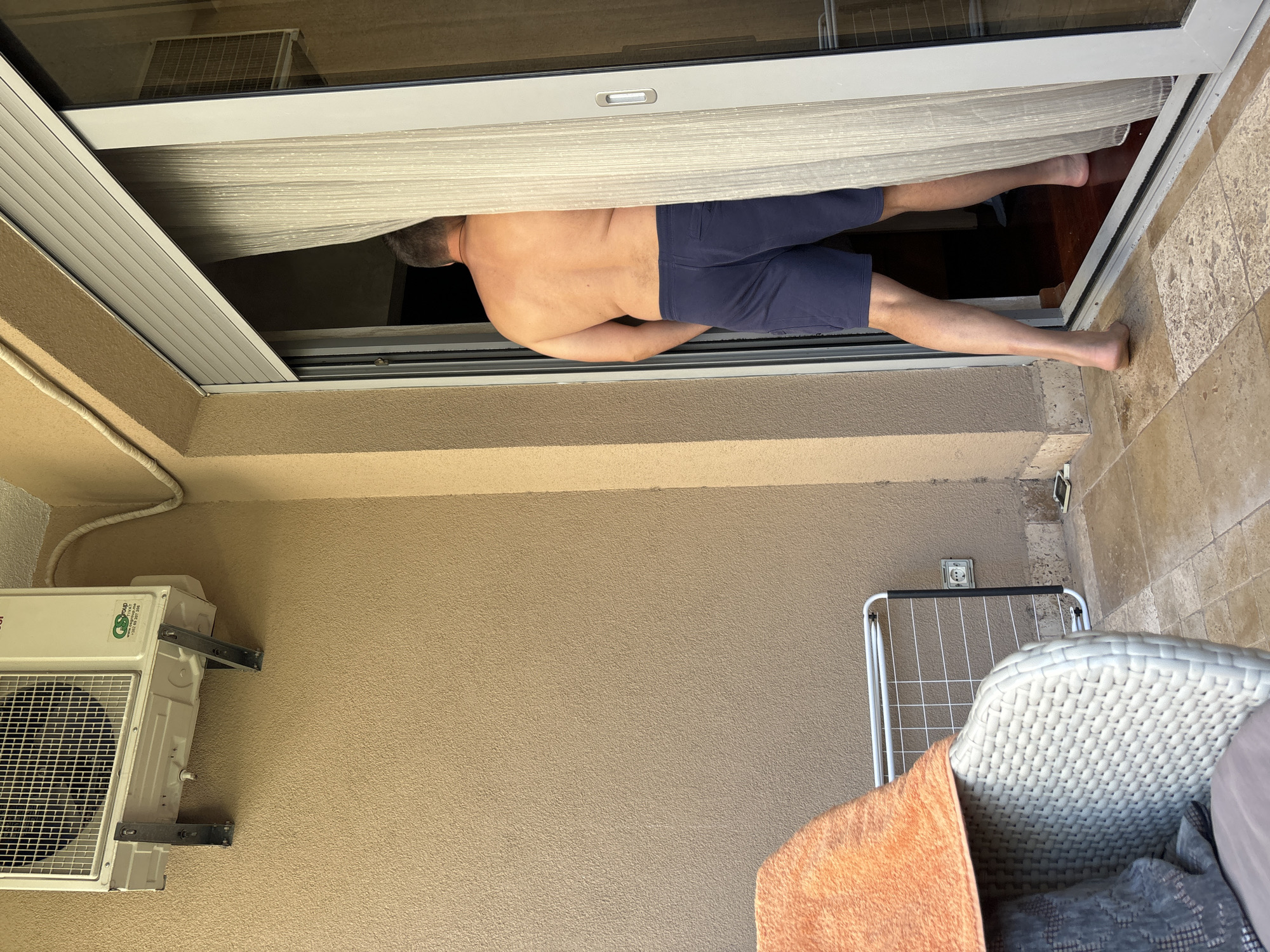 A man is semi-prone, torso and legs exposed, partially visible through a sliding glass door on a balcony. He is wearing dark blue shorts. The balcony has a tan-colored stucco wall on which an air conditioning unit is mounted. An electrical outlet and a clothes drying rack are also visible. A wicker chair with cushions and an orange towel are in the foreground. The location is likely in Budva, Montenegro.