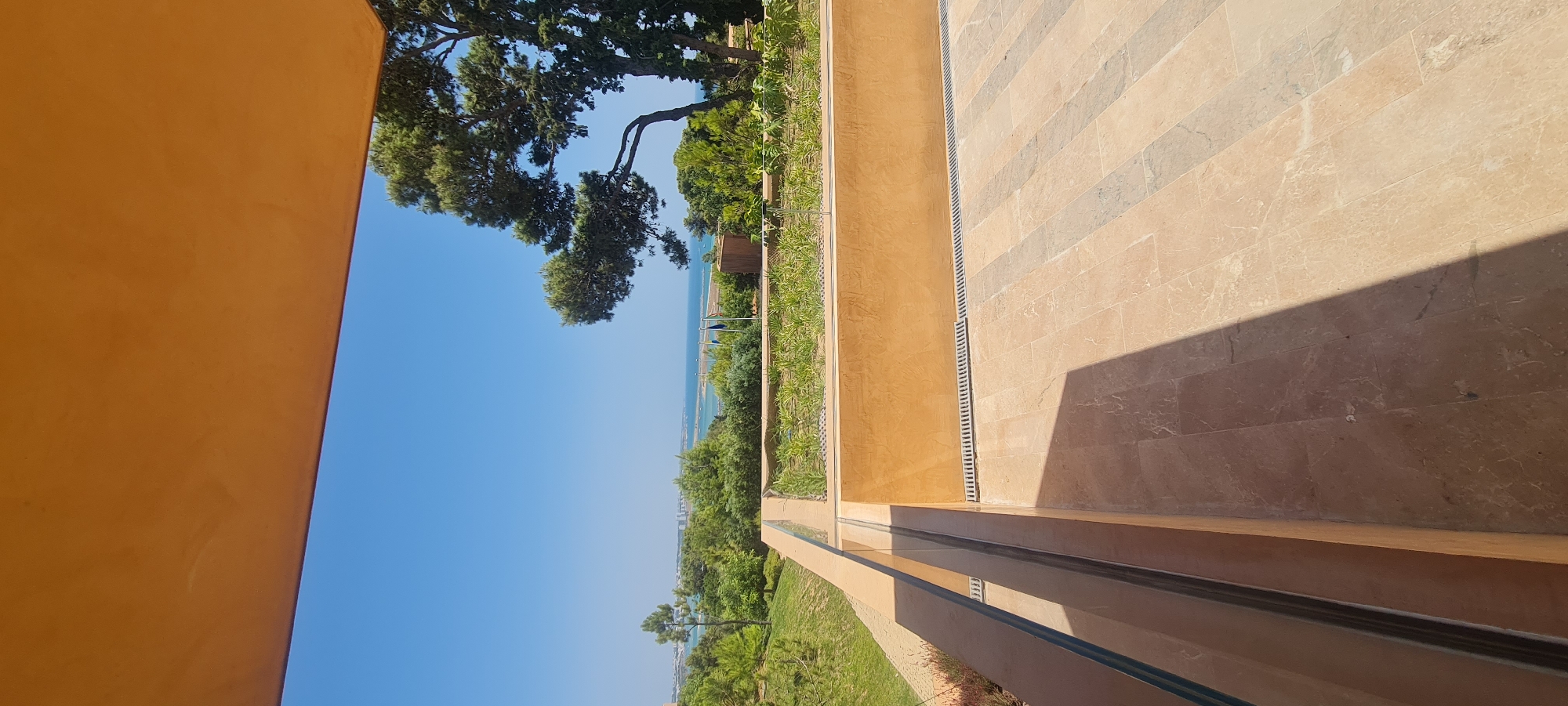 An outdoor terrace with marble flooring overlooks a bright blue sky, the Atlantic Ocean, and a green, hilly landscape with scattered buildings and trees. A large, textured orange overhang frames the top of the image. The terrace features a glass railing, a low, orange-hued wall with a drain grate, and a raised planter filled with green foliage. Shadows from the overhang fall across the patterned flooring. The scene appears to be located in Lagos, Portugal, likely a resort or residential area with views of the coast. There are no people visible in the image.