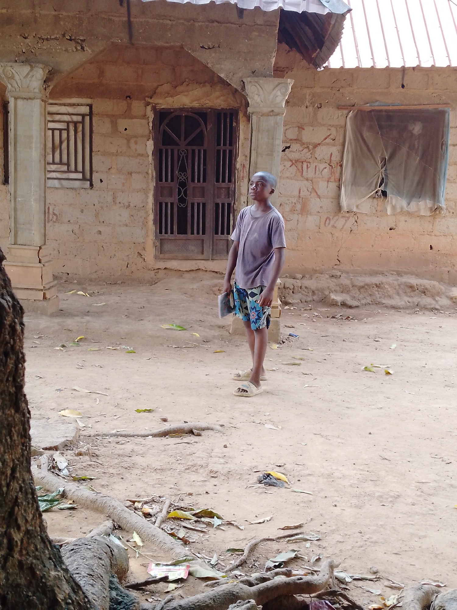 In Akwanga, Nigeria, a young boy stands in the dusty yard of a weathered, single-story building with a corrugated metal roof. He wears a t-shirt, patterned shorts, and sandals, and he holds a dark object in his left hand. The building features crumbling mud brick walls, a dark metal door, two windows (one covered with a tattered cloth), and decorative columns flanking the entrance. Tree roots and debris litter the ground.