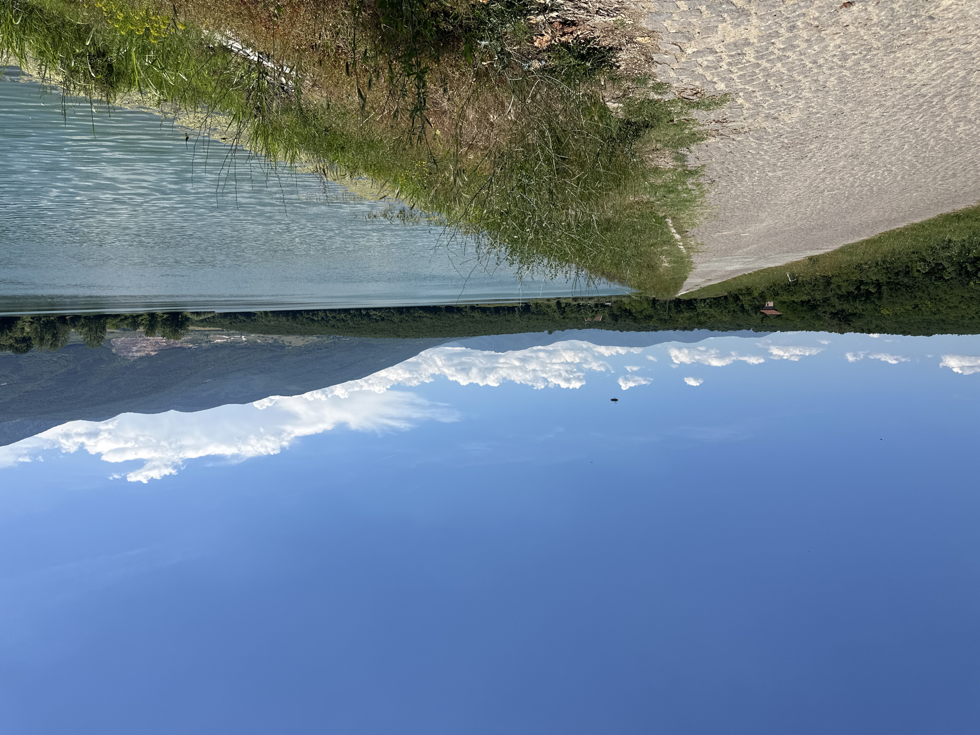 The image shows a paved pathway alongside a body of water under a clear blue sky with scattered clouds. The pathway is constructed with cobblestones and is bordered by dry grass and vegetation. To the left of the path, dense green trees form a line, with a small, reddish-brown building visible amongst them. The body of water, likely a lake or reservoir, is a calm, light blue with ripples on its surface. In the distance, rolling, tree-covered hills and mountains form the horizon. The sky is bright blue with a few wispy white clouds. There are no people visible in the image. The overall scene depicts a serene natural landscape with a man-made path. This location is within the city of Muldava, Bulgaria.