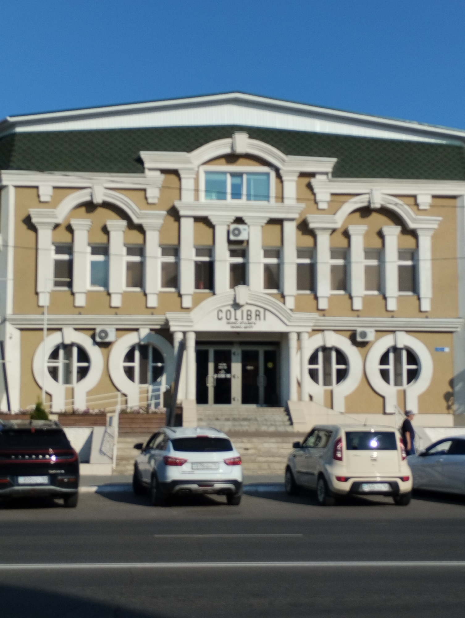A wide shot captures the facade of a beige, two-story building with ornate white trim and decorative columns. A dark green, pitched roof is visible above. The building has multiple windows, some arched and some rectangular. The text "COLIBRI" is prominently displayed above the entrance. Several cars are parked on the street in front of the building. The street is paved, and a clear blue sky is overhead. The scene appears to be a sunny day. Based on available information, this location is the "Colibri" building in Kostanay, Kazakhstan, located at 156 Talanov Street.