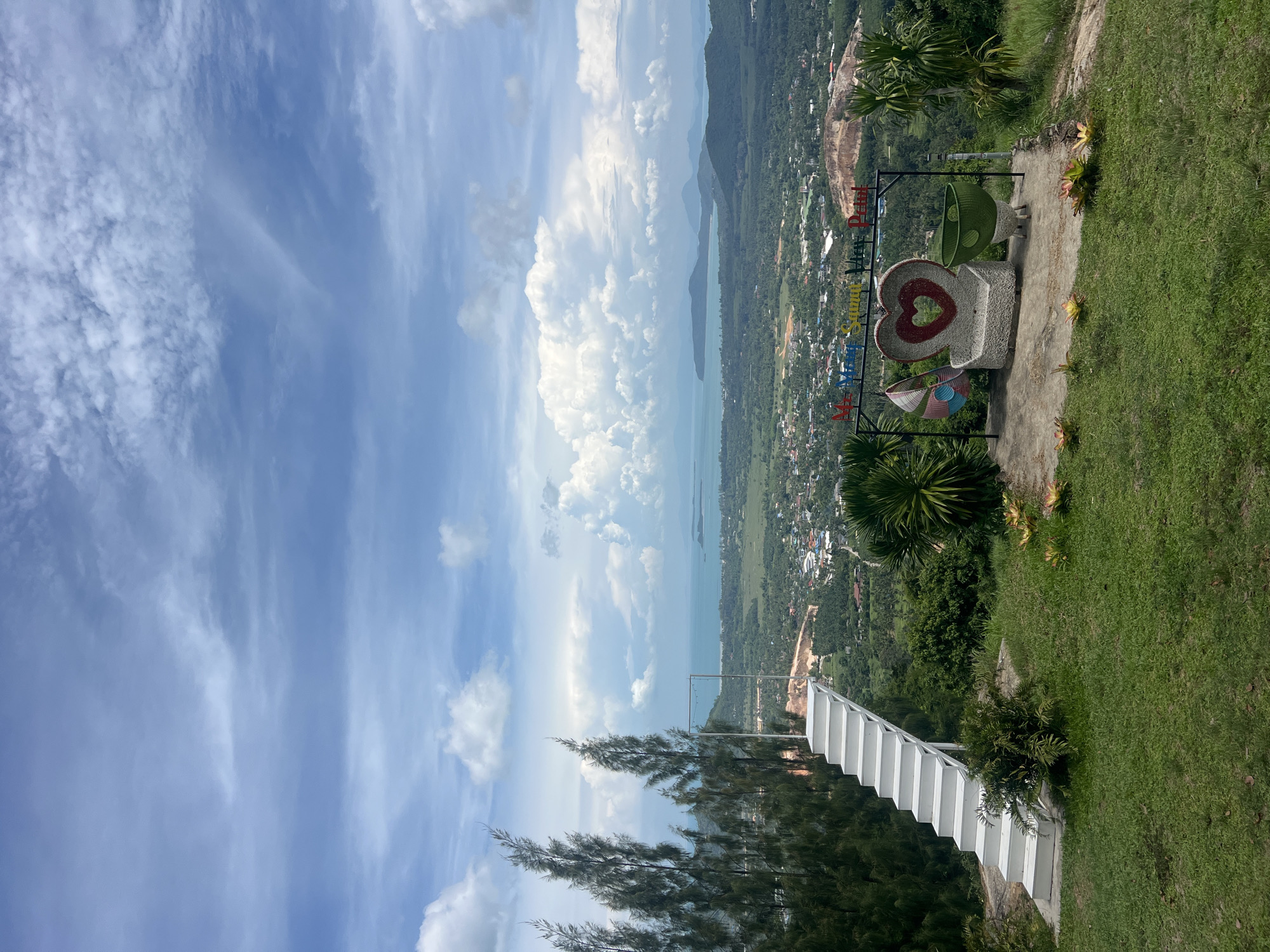 The image depicts a scenic viewpoint overlooking a coastal town and the ocean. In the foreground, there is a white staircase leading up to an observation deck. To the right of the staircase, there is a seating area with a bench shaped like a heart, adorned with colorful woven decorations. A sign above the seating area reads "Mr. View Samui View Point." The background shows a verdant landscape with a densely populated town, hills, and the sea. The sky is partly cloudy, with dramatic cumulus formations. The location is identified as Baan Thurian, Thailand. There are no people visible in the image.