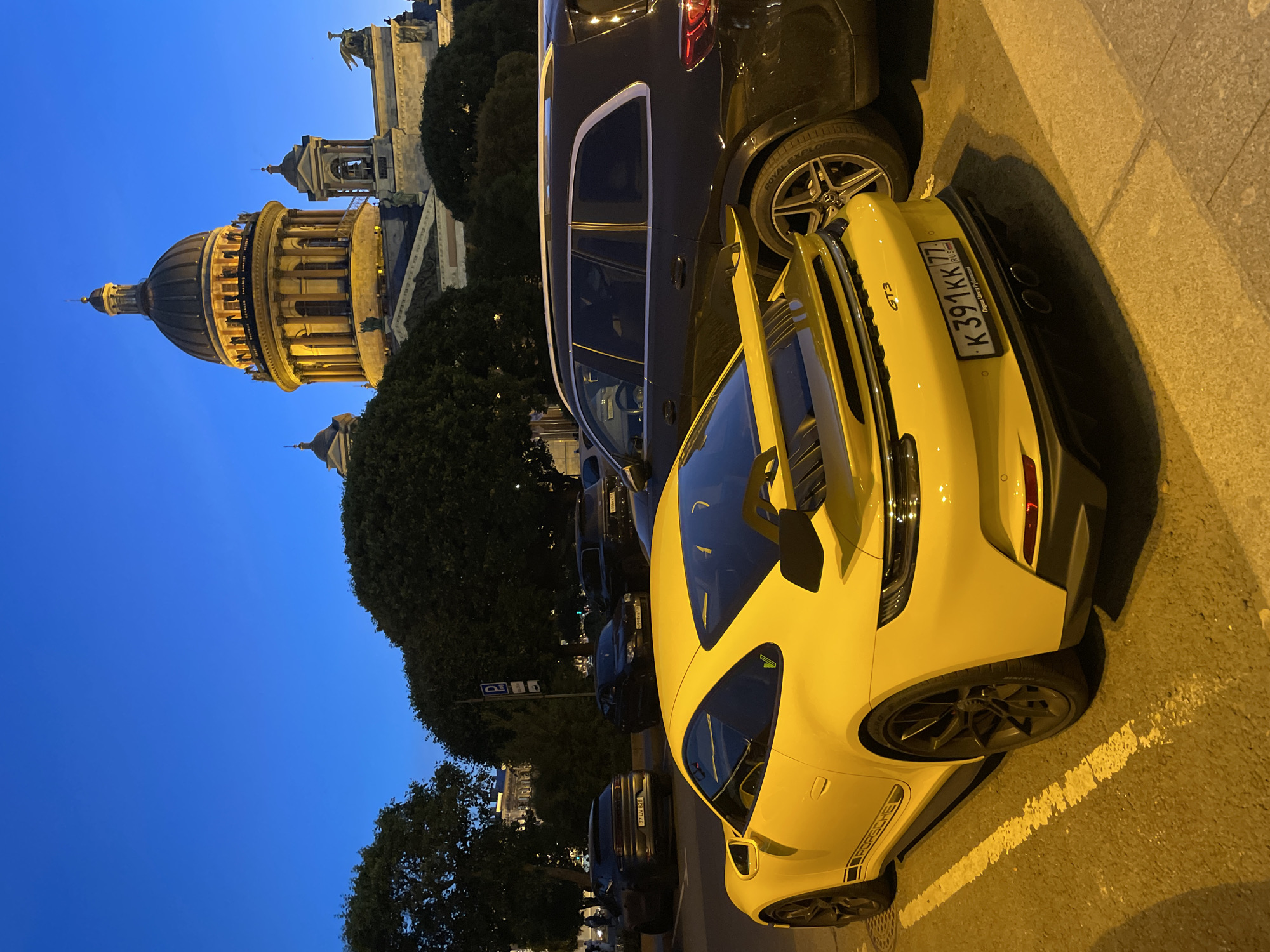 A yellow Porsche GT3 is parked in a lot in front of a large domed building, possibly St. Isaac's Cathedral in Saint Petersburg, Russia, as indicated by the license plate reading "K391KK 77 RUS". The car is positioned in the foreground, its rear end facing the viewer. Behind it, a dark-colored SUV is parked. The sky is a deep blue, suggesting it is either early morning or late evening. Several trees are visible around the parking area. The lighting is low, with the dome and some architectural elements of the building illuminated.