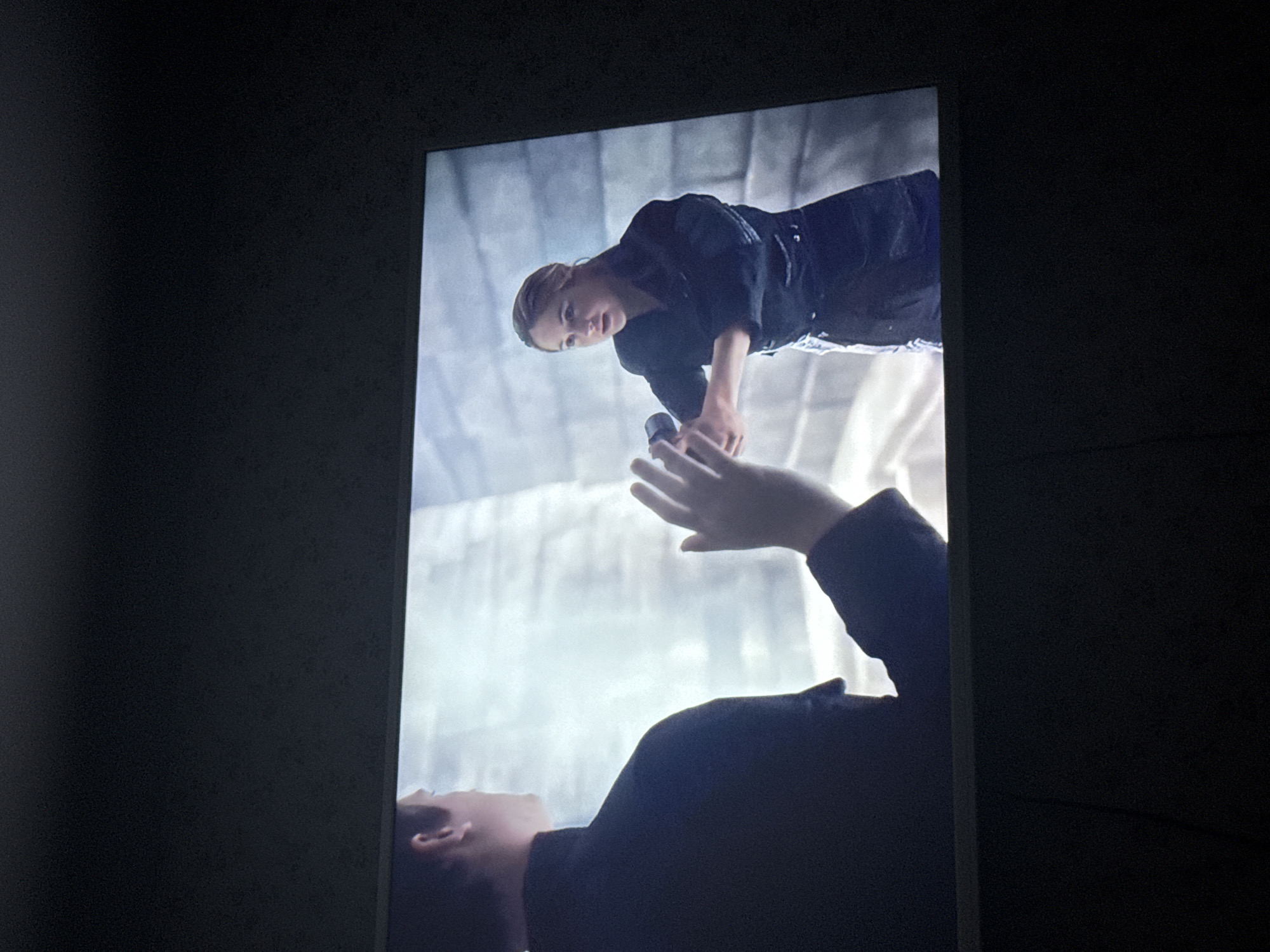 A television screen displays a scene of two figures: a woman in a dark outfit holding an object and a man looking up at her with his hand reaching up. The backdrop appears to be a concrete structure. The TV is mounted on a dark wall. The location is undetermined.