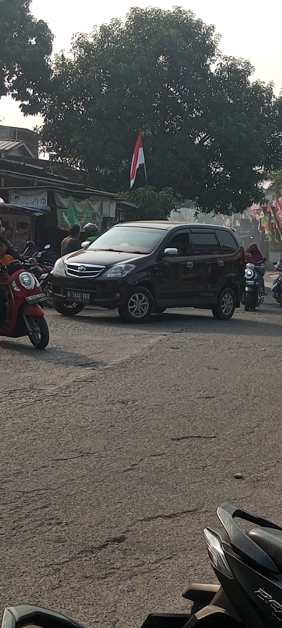 In Setu, Indonesia, a dark-colored minivan with license plate B 1846 FFV is seen on a paved road. The minivan has a red interior visible through the windows. The road surface is cracked and textured. To the left of the minivan, a red scooter is parked, also with a license plate. Behind the minivan, a person wearing a red headscarf is riding a motorcycle. Several other motorcycles are visible in the background, some with riders. A row of buildings with awnings and signs lines the left side of the road. A red, white, and blue flag is prominently displayed on a flagpole behind the buildings, silhouetted against a bright sky. Lush green trees with dense foliage frame the upper portion of the image. Another motorcycle is partially visible in the foreground, its rear end and seat in focus.