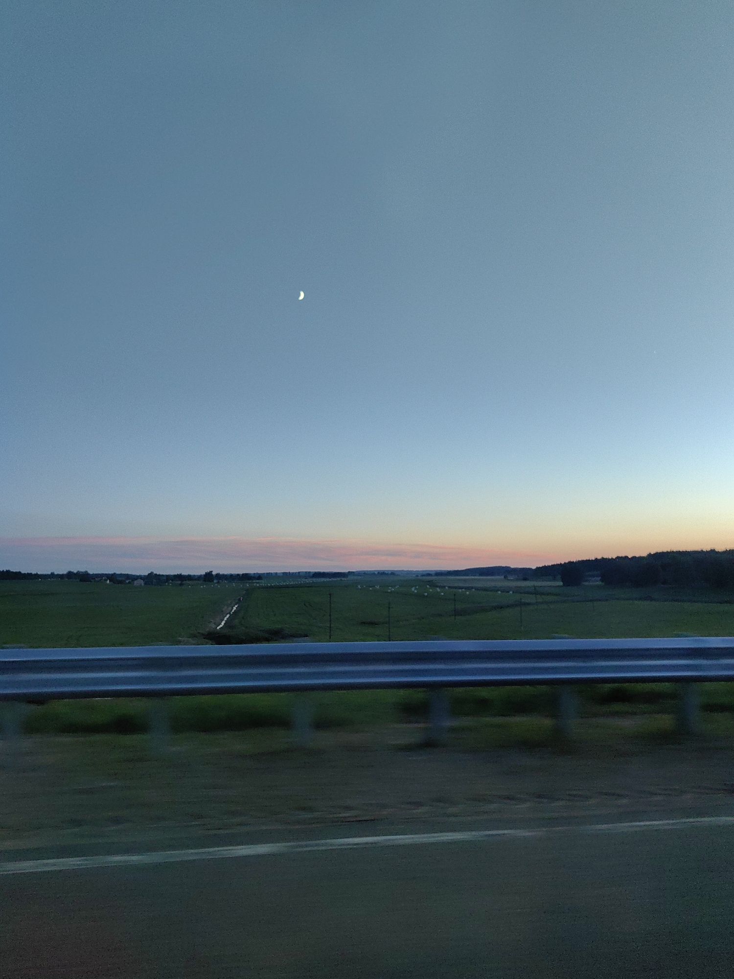 This image depicts a rural landscape at dusk, likely in the Faniapal Rural Council area of Belarus. The foreground features a metal guardrail, common along roadsides, with a blurred, dark-colored road surface below it, suggesting the photo was taken from a moving vehicle.
Beyond the guardrail, a vast, green field stretches out, appearing to be agricultural land. A thin, winding ditch or small stream can be seen cutting through the field. In the distance, a cluster of buildings, possibly a village or farm settlement, is visible. The horizon is marked by a line of trees, creating a silhouette against the fading light. The sky is a gradient of colors, transitioning from a pale blue at the top to soft pinks and oranges near the horizon, indicative of sunset or twilight. A small, crescent moon is faintly visible in the upper left portion of the sky. The overall mood is serene and peaceful, characteristic of the end of the day in a rural setting. There is no visible text in the image.