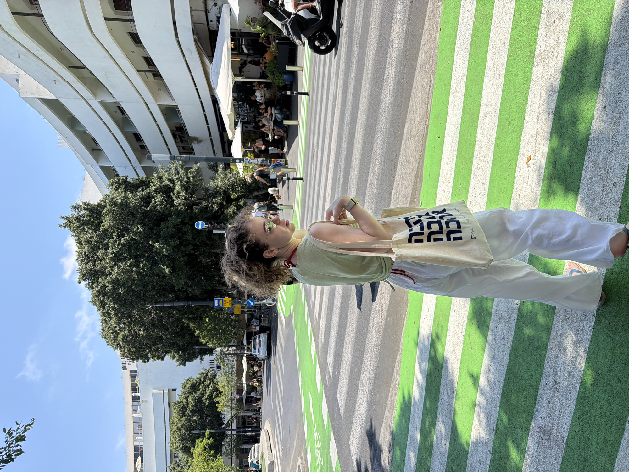 The image captures a scene on a Tel-Aviv street. A young woman with curly hair is standing on a crosswalk with green and white stripes. She is wearing a light green top, white pants, and sunglasses, and carrying a tote bag. The street setting includes a restaurant with outdoor seating, a scooter parked nearby, trees, a bus stop, and other vehicles in the background. A tall, white building stands in the background on the left.