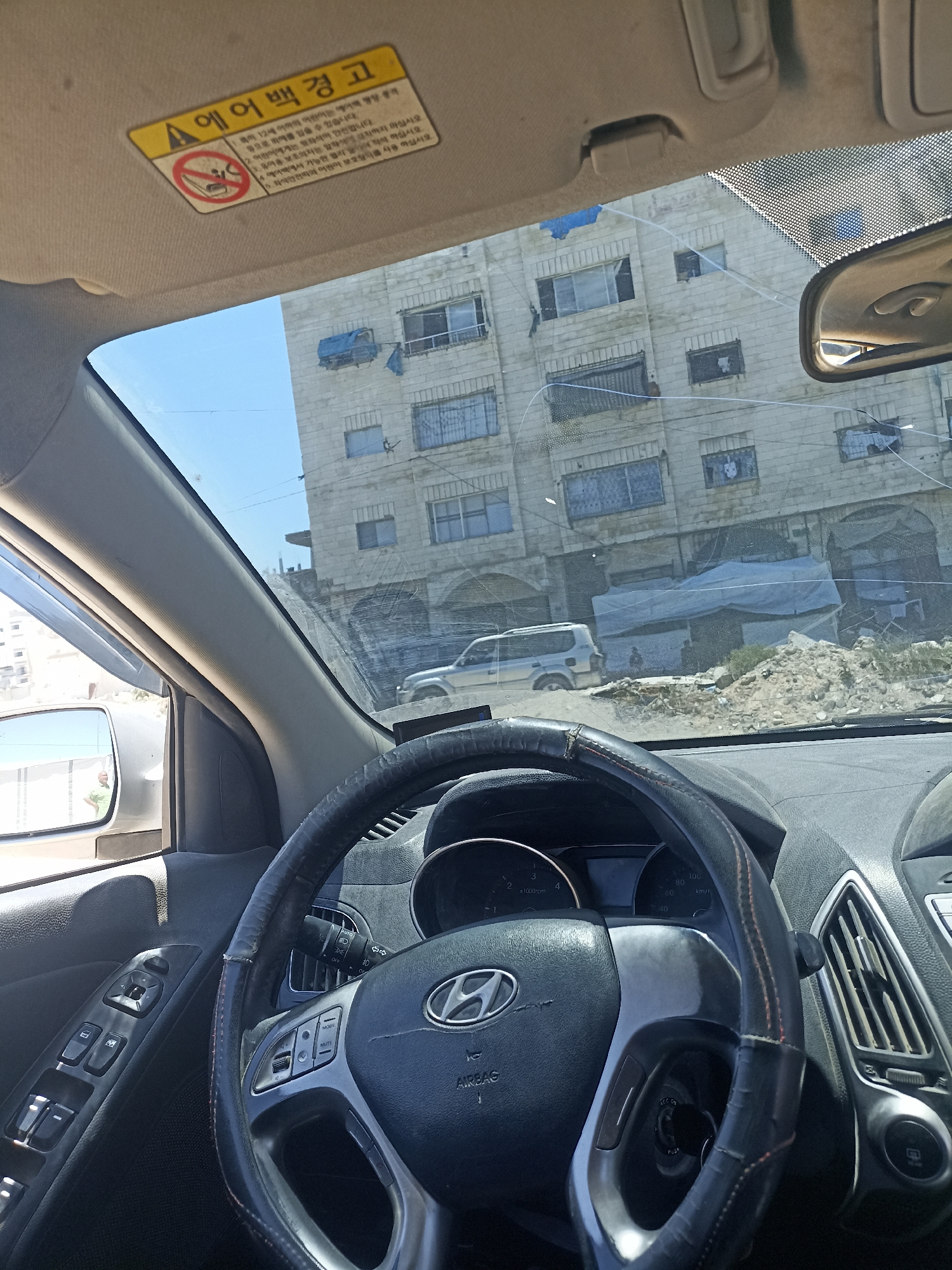 The image is taken from inside a car, looking out through the windshield. The subjects are the car's interior, including the steering wheel, dashboard with visible instruments like a speedometer and tachometer, and the driver's side door controls. Outside, the scene is a building with multiple floors and windows, appearing somewhat run-down, with debris and makeshift structures in front of it. A silver SUV is parked in front of the building. The sky is clear and blue, suggesting daylight, likely late morning or early afternoon. There is a yellow warning sticker on the car's sun visor with Korean text and a pictogram indicating an airbag warning for children. The text translates to "Airbag Warning" and provides instructions related to children and airbags. The overall impression of the scene is one of a moderately populated urban or semi-urban area, with signs of potential hardship or less-than-ideal living conditions. No people are clearly visible in the external scene, though a figure can be glimpsed in the side mirror. The image captures a moment of stillness, possibly during a pause in driving.