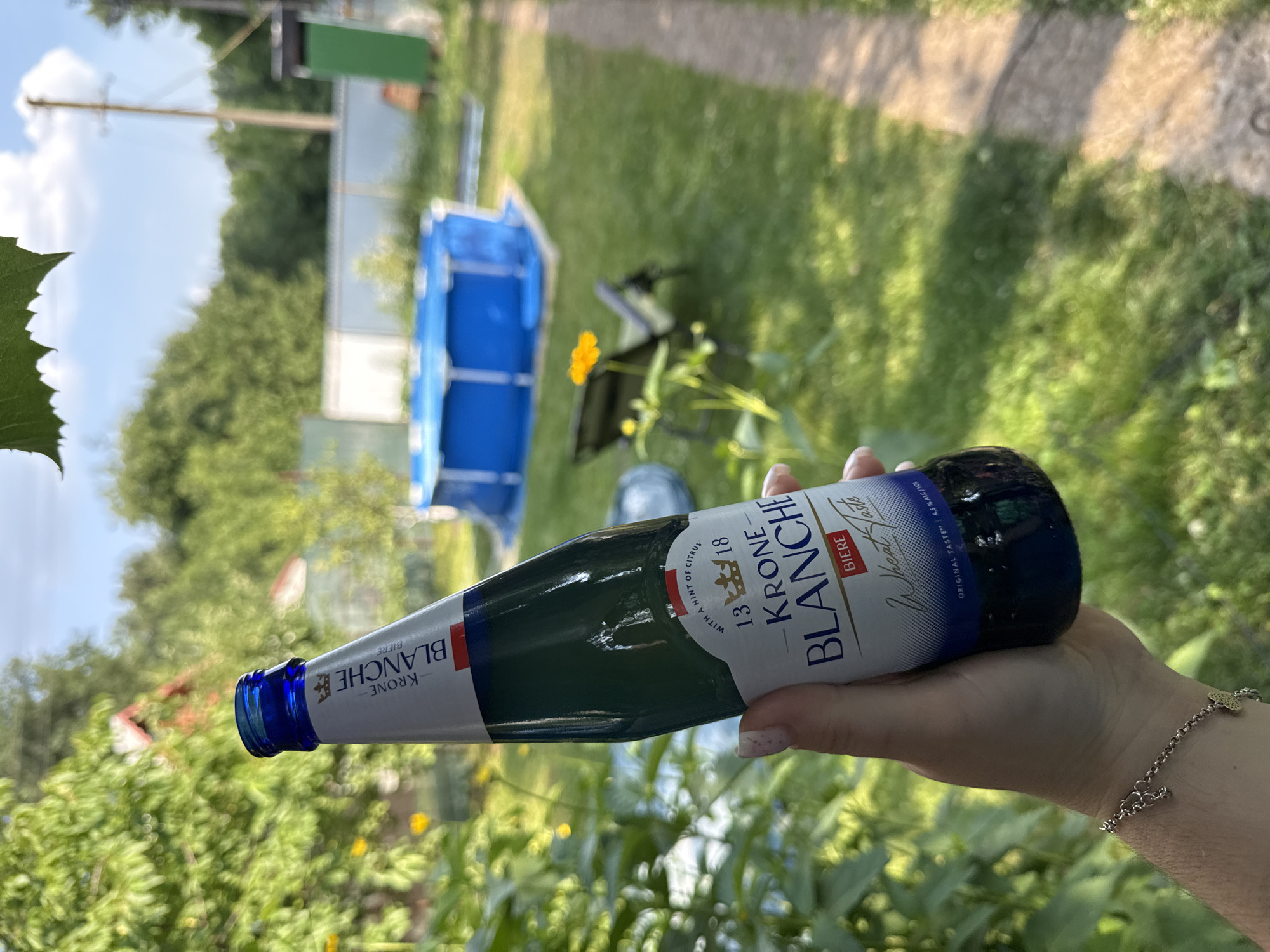 A person's hand, adorned with a delicate bracelet, holds a glass bottle of Krone Blanche Biere. The bottle, which appears to contain a pale liquid, has a label with text including "KRONE BLANCHE BIERE," "Wheat Fasts," and "4.5% ALC/VOL." The label also mentions "WITH A HINT OF CITRUS." The background suggests an outdoor setting, likely a backyard, with lush green grass and trees. A blue inflatable pool is visible in the distance, along with a path or walkway. The lighting suggests it is daytime, possibly a sunny day given the bright green foliage. The image is oriented sideways, with the bottle held horizontally.