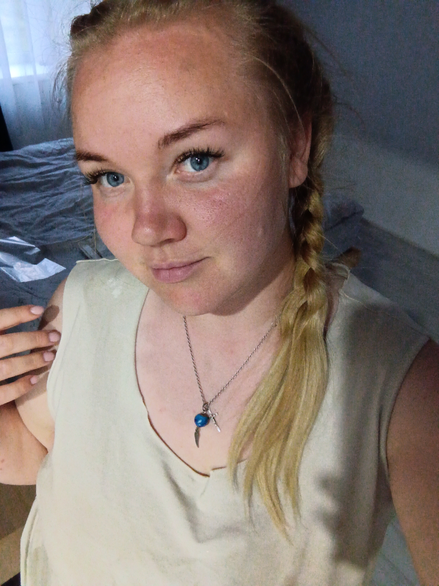 A woman with blond hair, styled in a braid on one side and pulled back from her face, is looking slightly off-center to the left. She has fair skin, blue eyes, and a natural complexion. She is wearing a light-colored sleeveless top and a silver necklace with a pendant. The background is a room with a bed partially visible. There are no discernible landmarks to determine if the image was taken in Фоки, Russia.