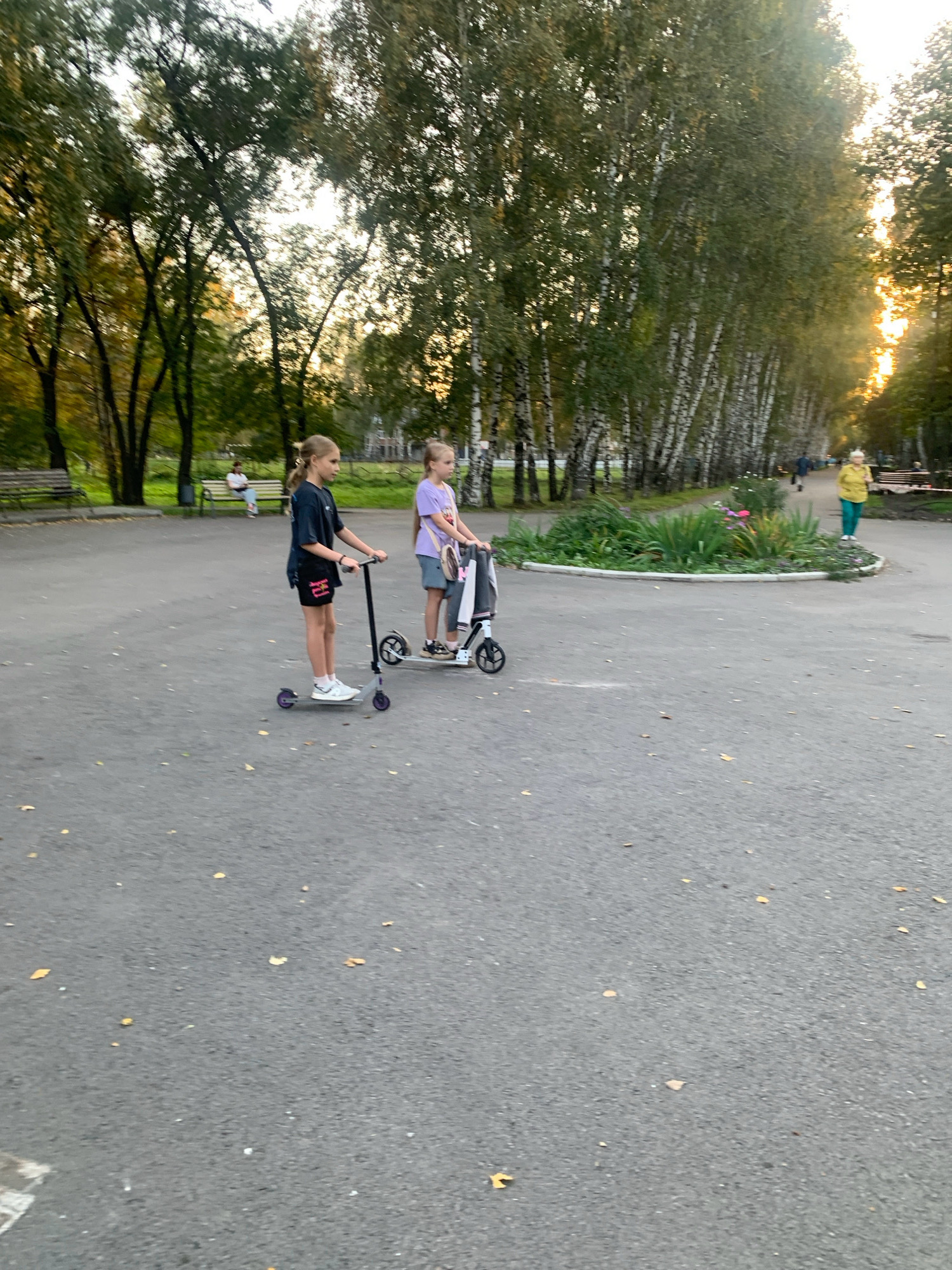 Two young girls are riding scooters on an asphalt path in a park. The girl on the left is wearing a dark t-shirt and shorts, and the girl on the right is wearing a purple t-shirt and shorts. They are both looking forward as they ride. The path is surrounded by trees, and there are benches and a flower bed in the background. The sun is setting, casting a warm glow on the scene. The location is the park in Prokopyevsk, Russia.