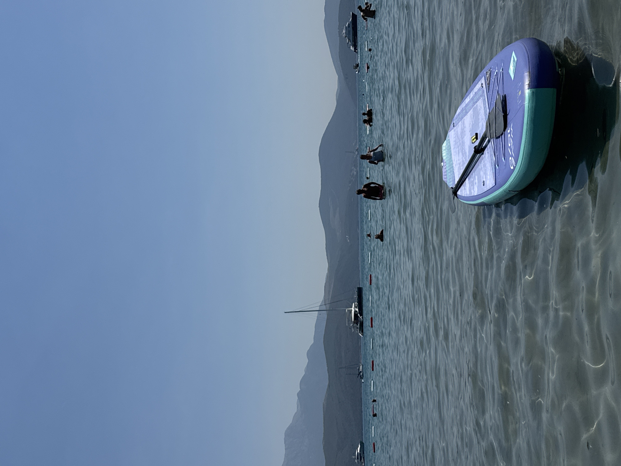 In Radovići, Montenegro, on a bright, sunny day, a paddleboard is floating in the shallow, clear water near the shore. The paddleboard is a vibrant blue and teal color, with a black paddle resting on top. In the background, several people are visible in the water, some wading and others swimming. Beyond the swimmers, a row of buoys marks a designated swimming area. Further out in the bay, various boats are anchored, including sailboats and a large yacht. The scene is framed by rolling, green hills and mountains under a clear blue sky.