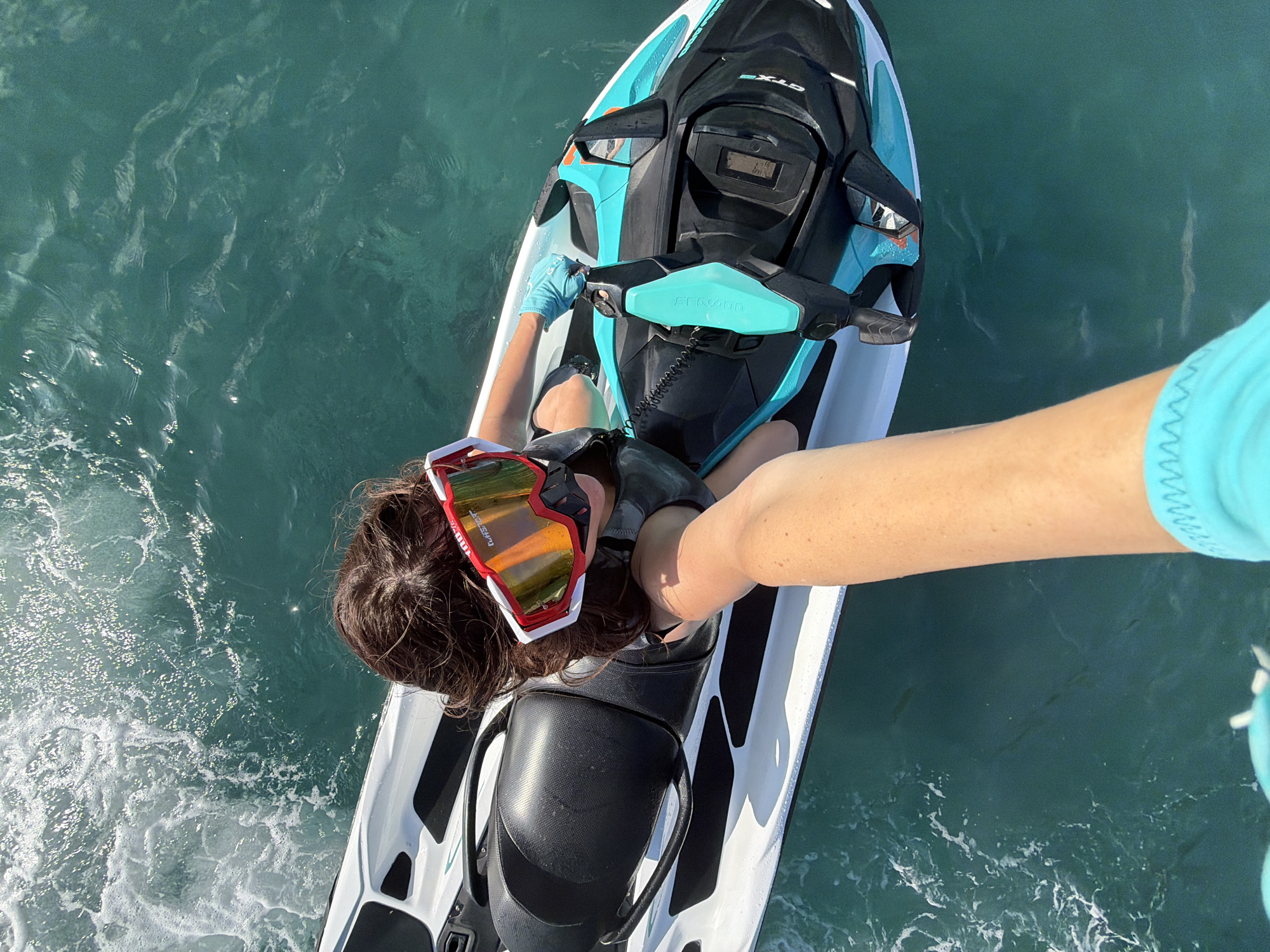 A woman with dark hair wearing red and white goggles and a black life vest is sitting on a white and teal jet ski. She is wearing a light blue glove on her right hand and holding the handlebars of the jet ski. The jet ski is on a body of blue-green water with white foam on the left side. The water is choppy and the sun is reflecting off the surface. The image is taken from a high angle, looking down at the woman and the jet ski. The background is out of focus and shows more water. The location is Nusa Penida, Indonesia, a tropical island.