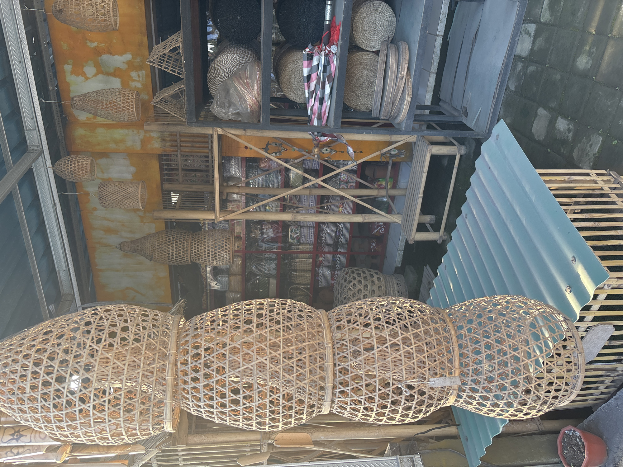 This image captures a shop scene in Ungasan, Indonesia, displaying a variety of handcrafted items. The focal point is a series of large, woven baskets, constructed from light-colored bamboo, strung together horizontally. These are suspended in the foreground, casting shadows onto the background.
Further back, the shop's interior is visible, revealing shelves stacked with more baskets of different shapes and sizes. Some are round and tightly woven, others are taller and more conical. A display of textiles and other decorative items lines the shelves.
The background features architectural elements, including walls and roofs. A section of corrugated metal roofing is visible, while other parts are painted in yellow. The lighting suggests an outdoor setting.